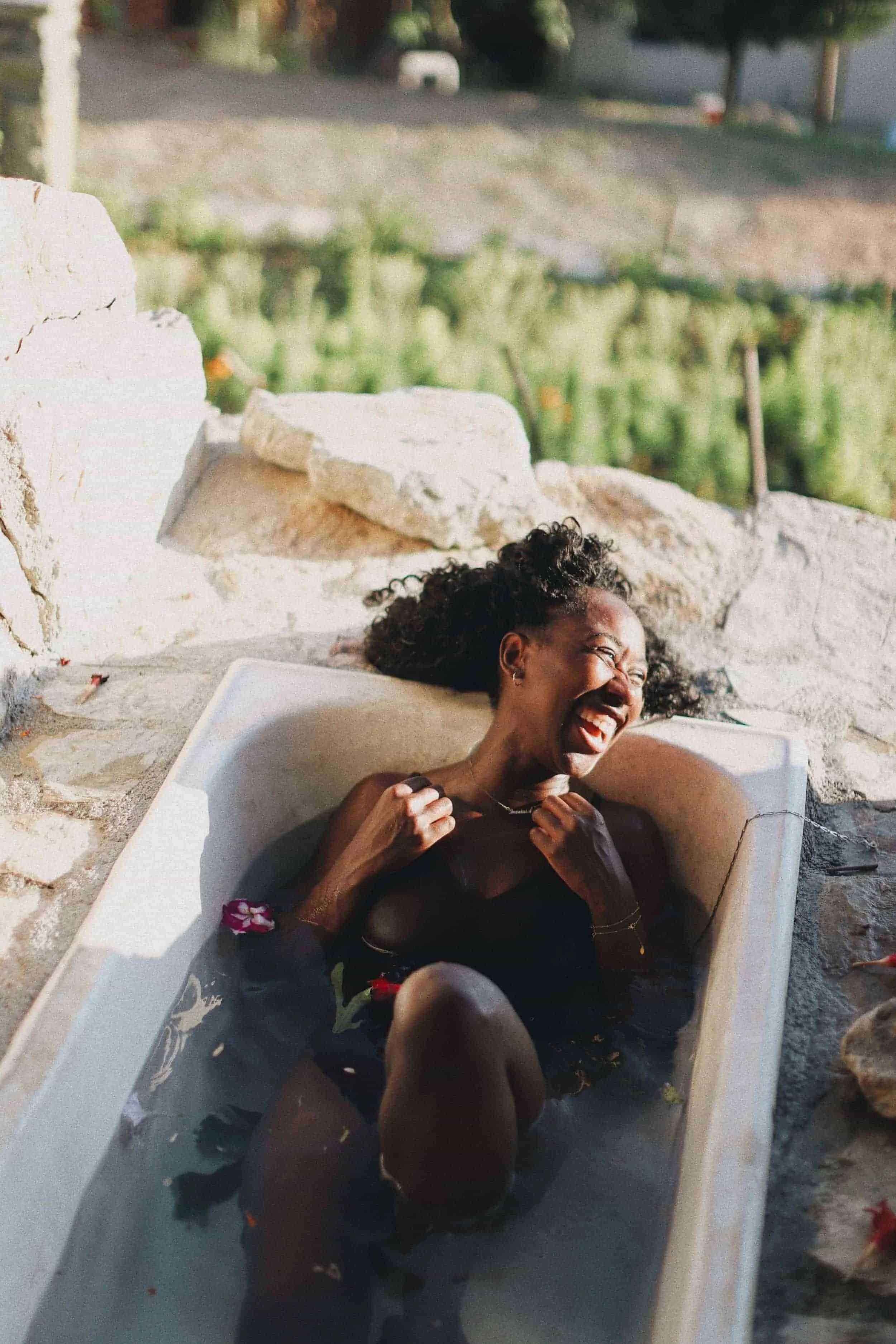 A woman laughing and relaxing in an outdoor stone bathtub filled with water and flower petals.