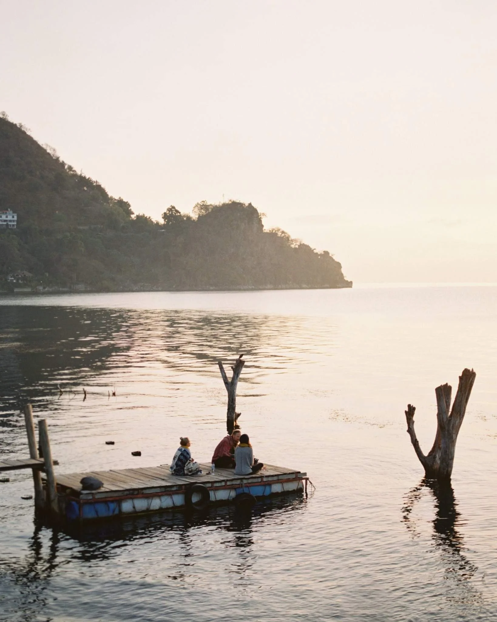 Three people sitting on a small wooden raft in a calm lake during sunset, with two leafless trees protruding from the water and a forested hill in the background.