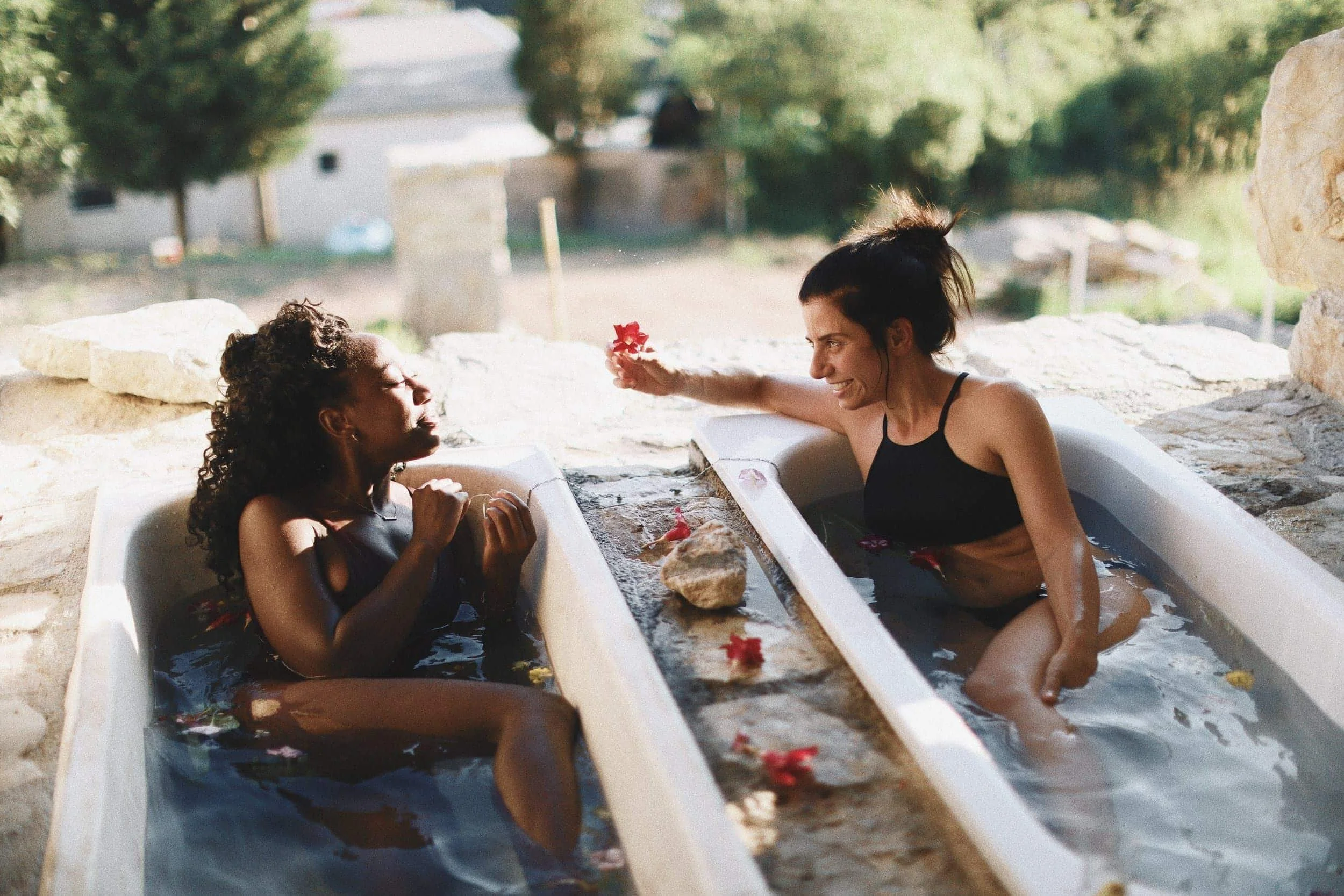 Two women relaxing in outdoor bathtubs, one with curly hair and the other with dark hair in a bun, smiling and talking while surrounded by waterfalls and rocks.