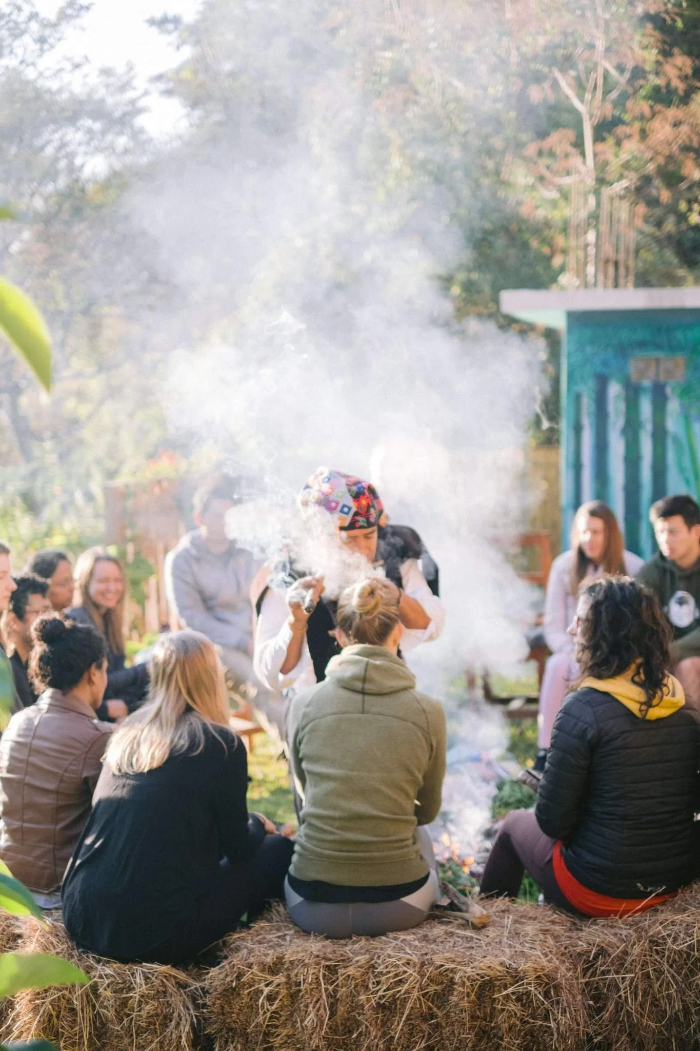 Group of people sitting outdoors on hay bales, watching a woman in a colorful headscarf performing a demonstration with smoke.