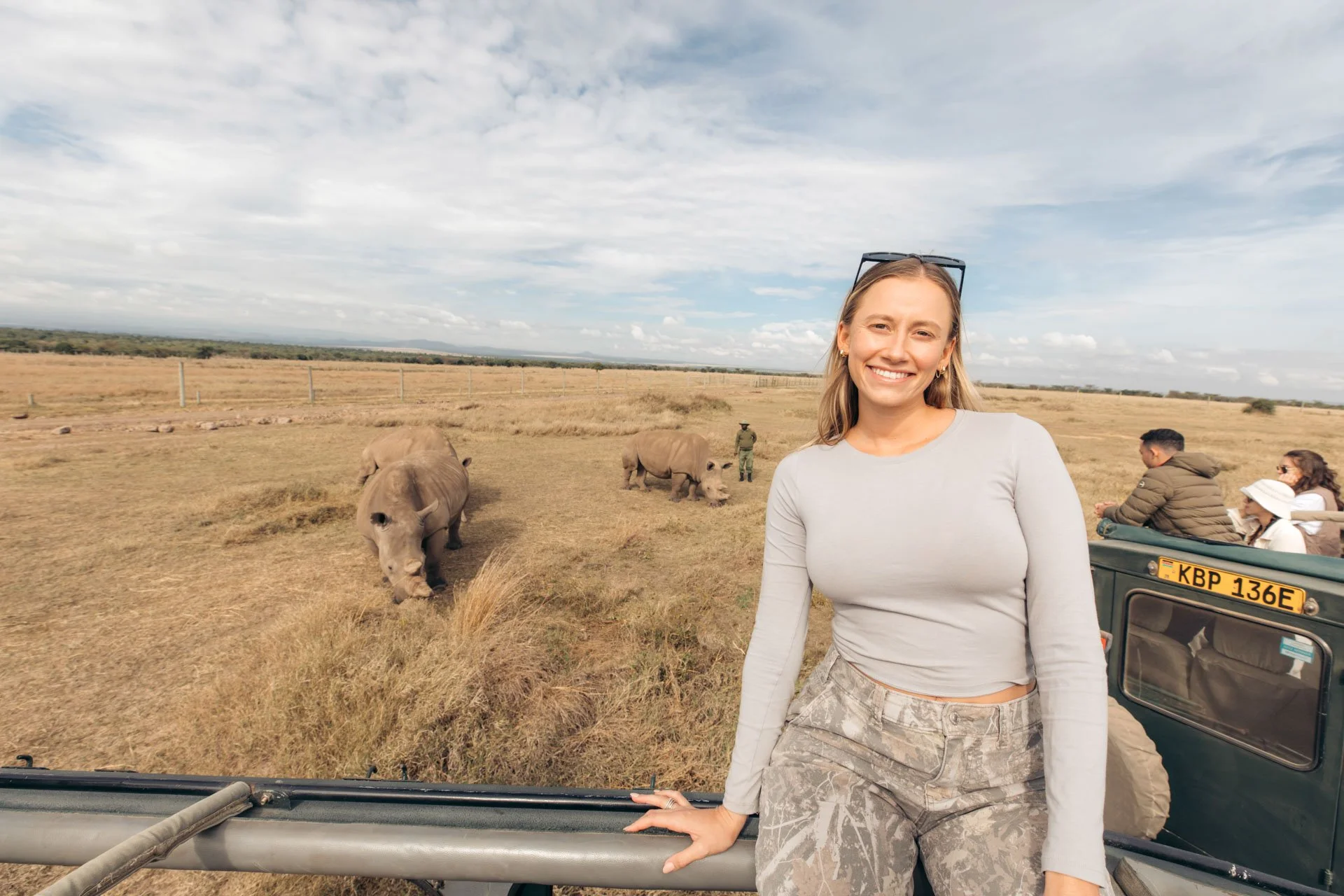 Smiling woman in long sleeve shirt and camouflage pants standing in open vehicle with safari view, safari animals grazing in the background, and a scenic sky.