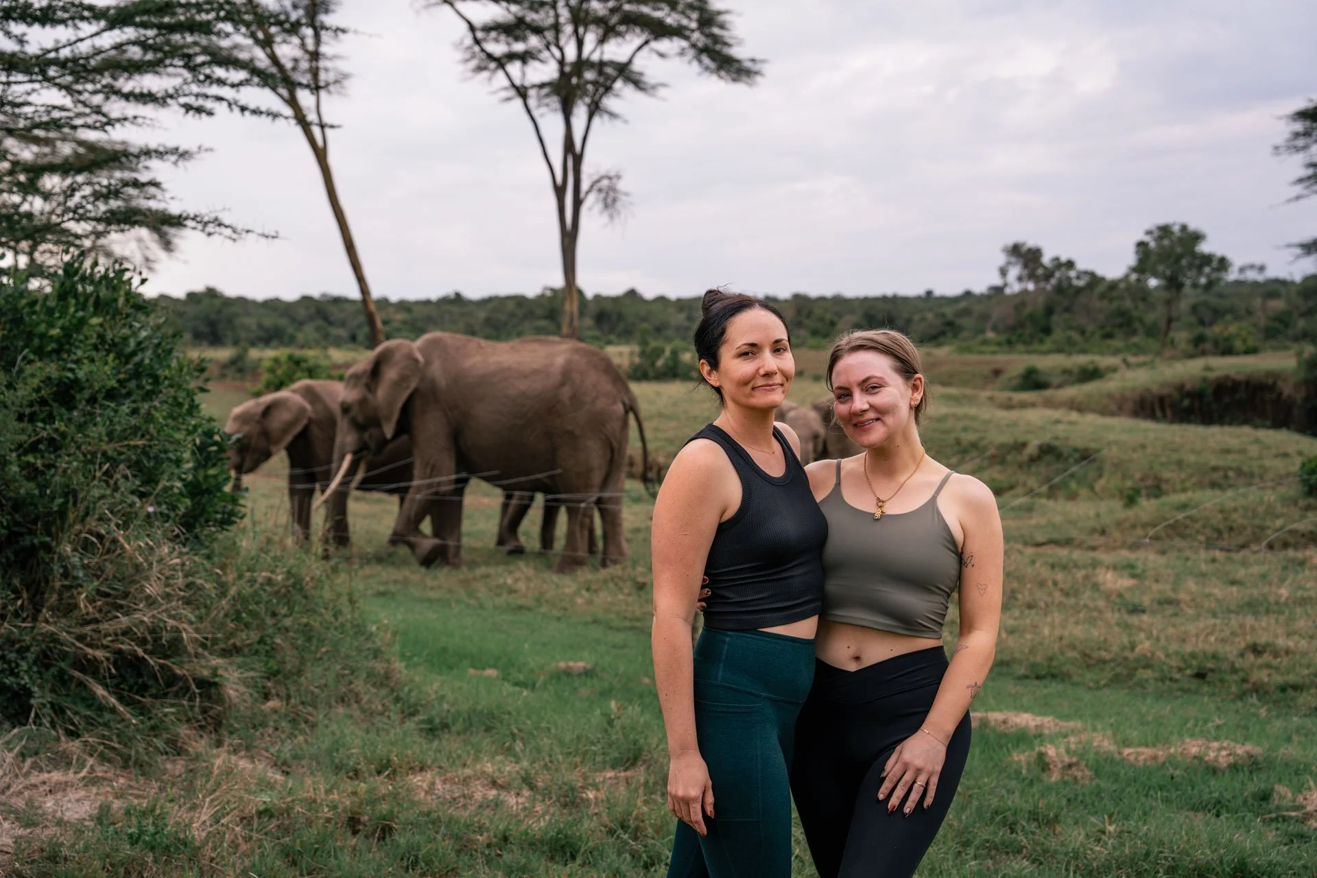 Two women standing outdoors with elephants in the background.