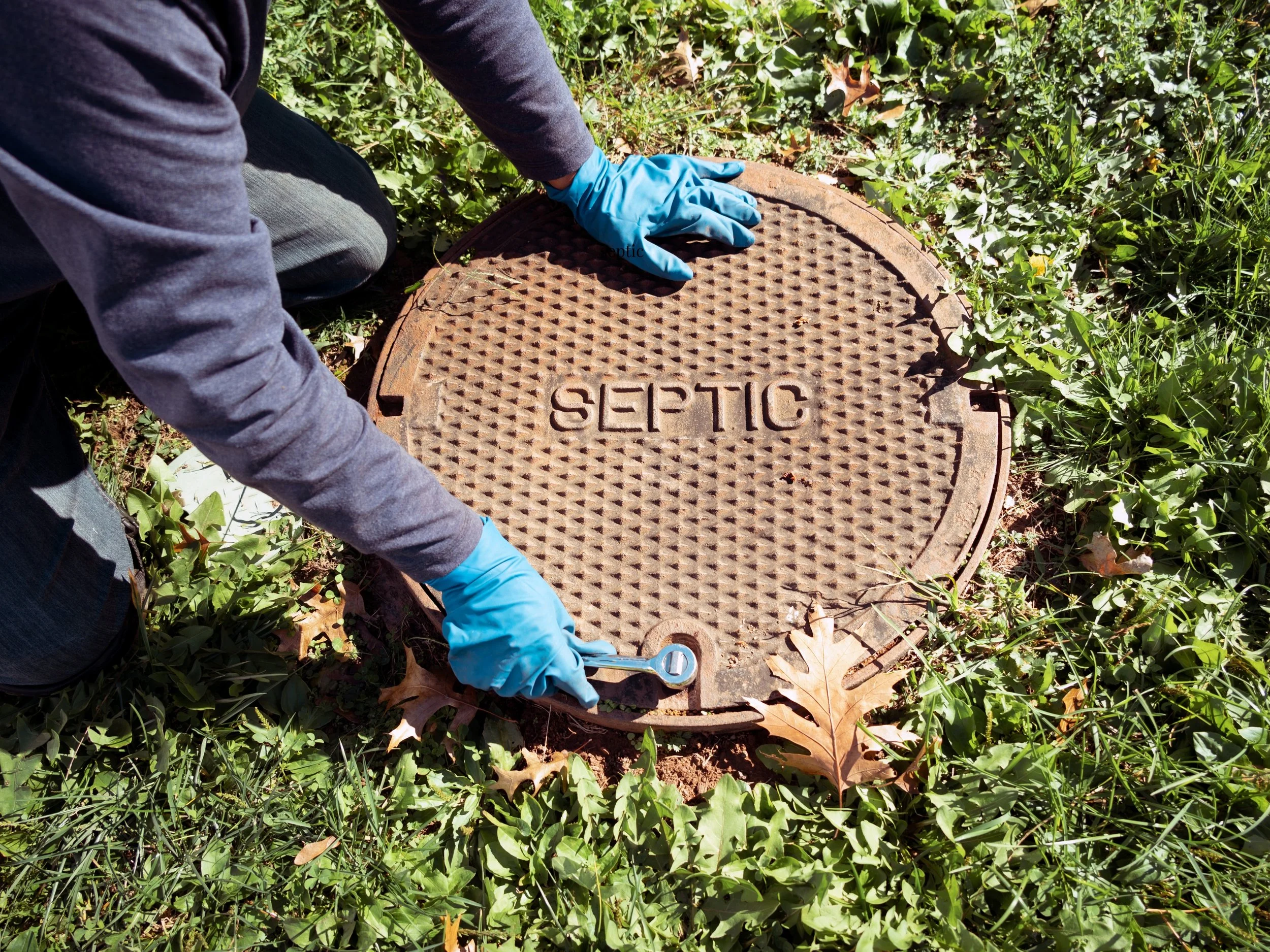 Person in blue gloves using a wrench on a septic cover in grassy area.