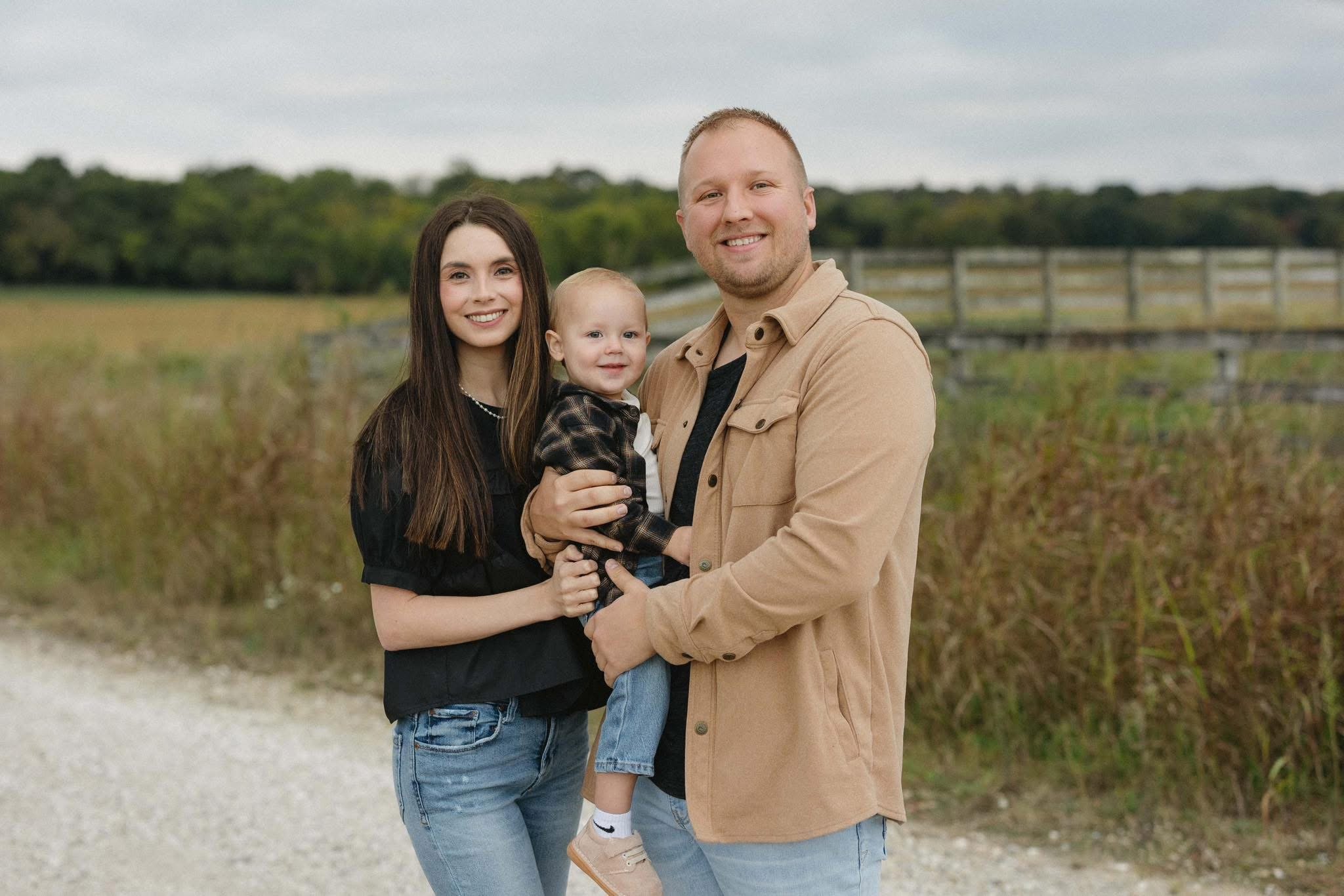 A family of three standing outdoors on a gravel path with a field and trees in the background. The woman has long brown hair and is wearing a black top, the man has short blond hair and is wearing a tan jacket, and the young child is being held by the man, wearing a plaid shirt and light-colored shoes.
