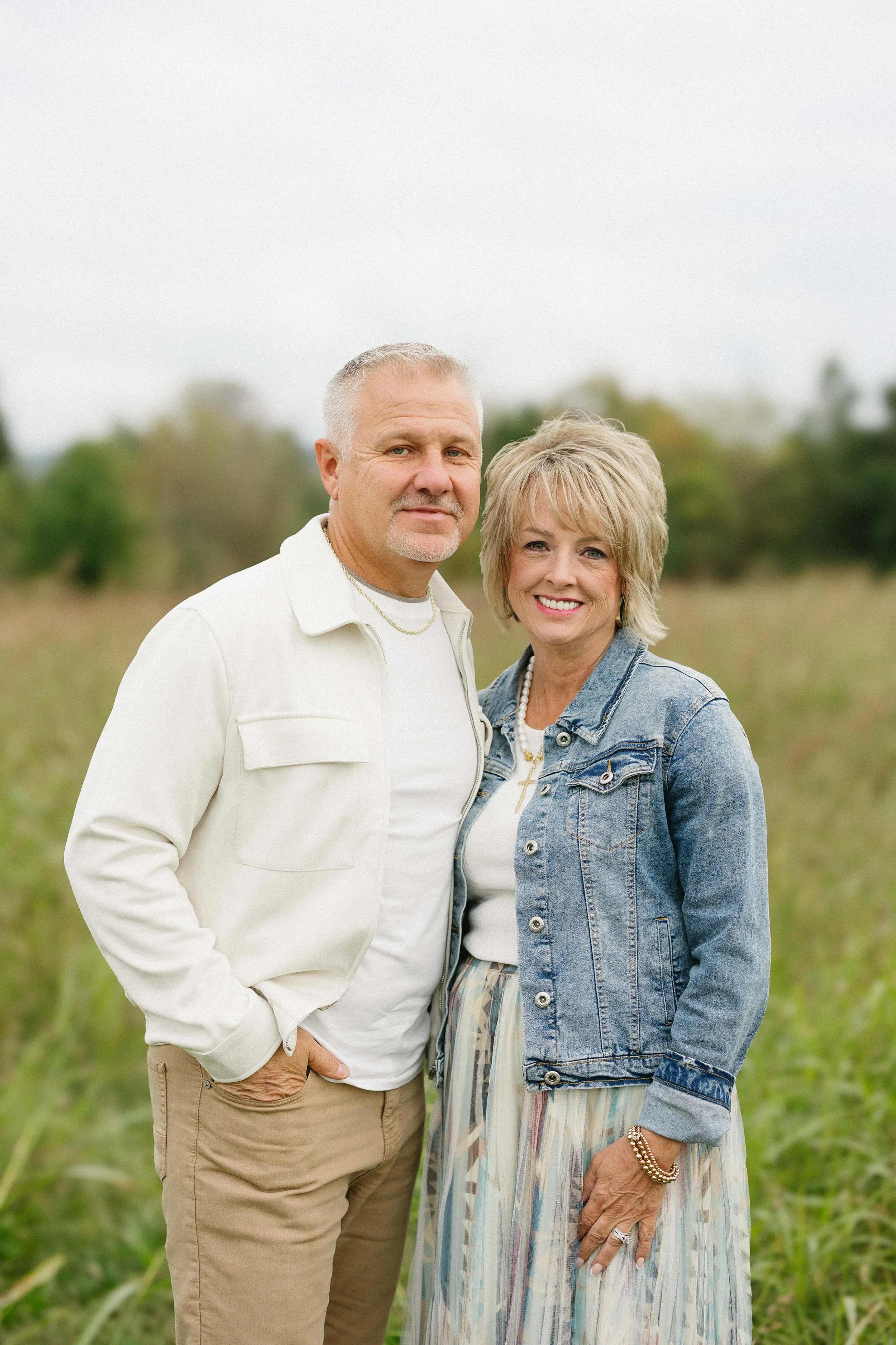 A smiling middle-aged couple hugging in an open grassy field with trees in the background, during sunset.