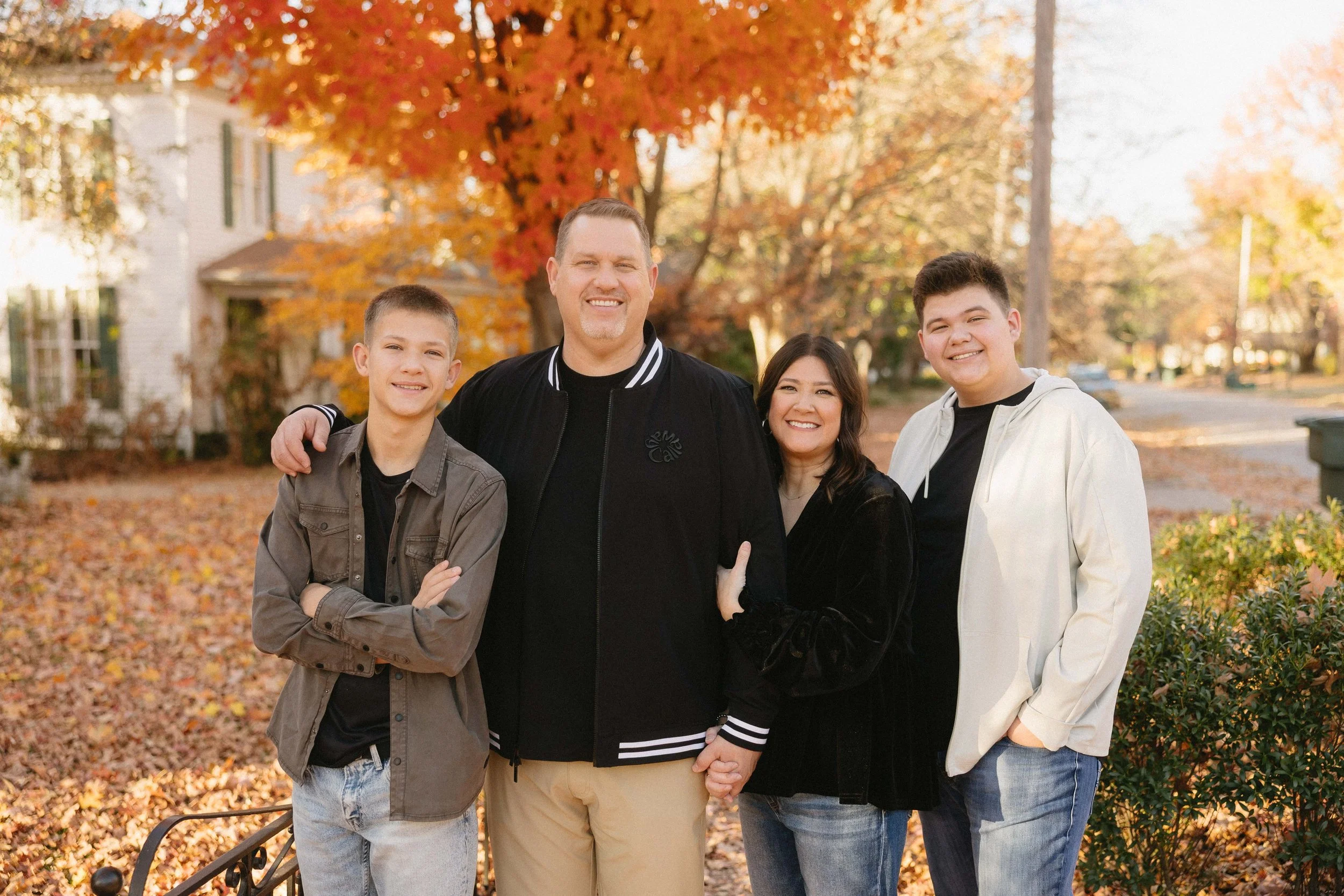 A family of five standing outdoors in autumn, with fall leaves and trees in the background. The family members are smiling and posing together, with two boys and two adults, one man and one woman.