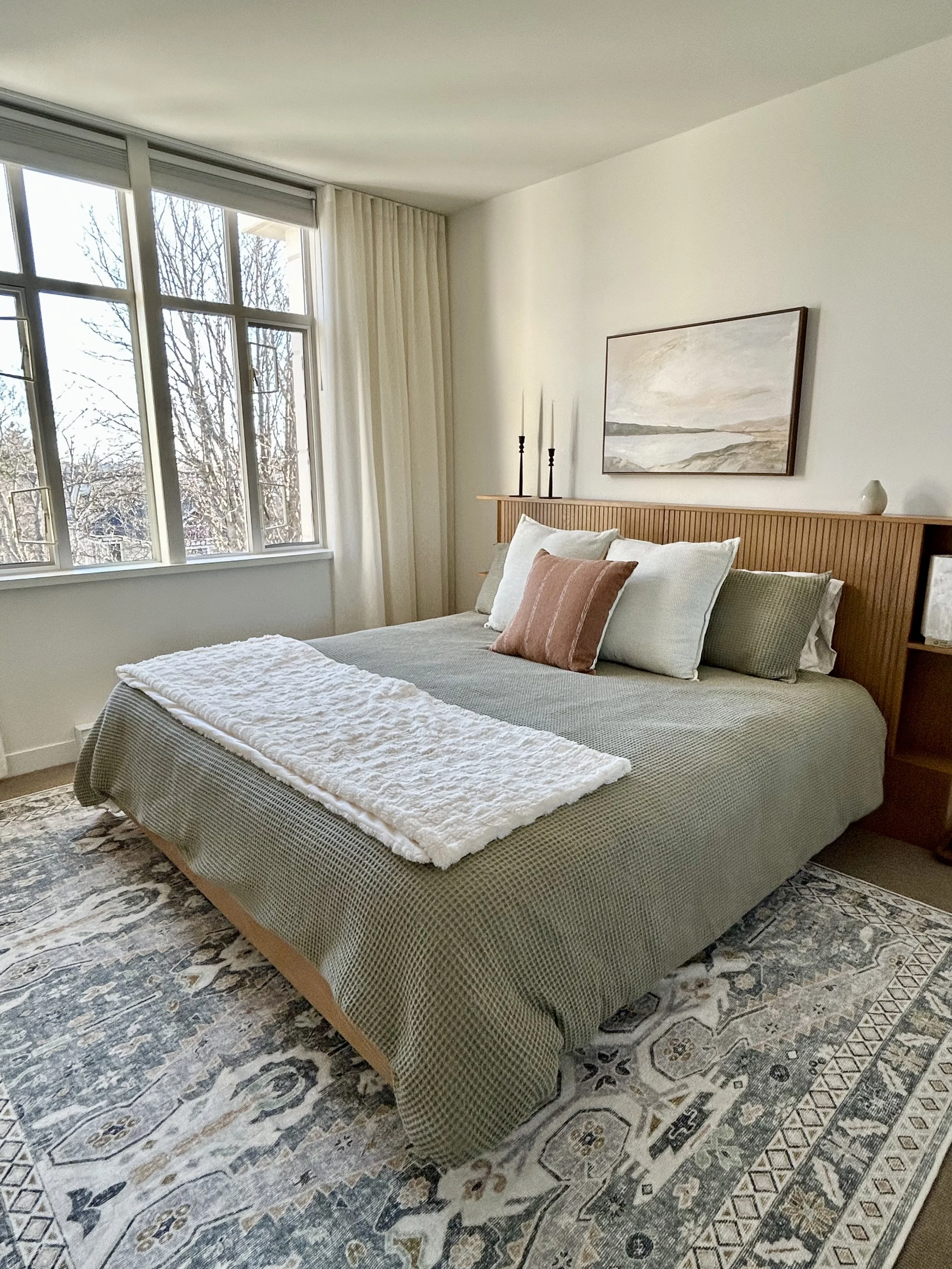 A bedroom with a large bed, green quilt, white pillows, pink pillow, and a white throw blanket. There is a wooden headboard, a framed landscape painting, and a window letting in natural light with curtains. An ornate rug covers the floor.