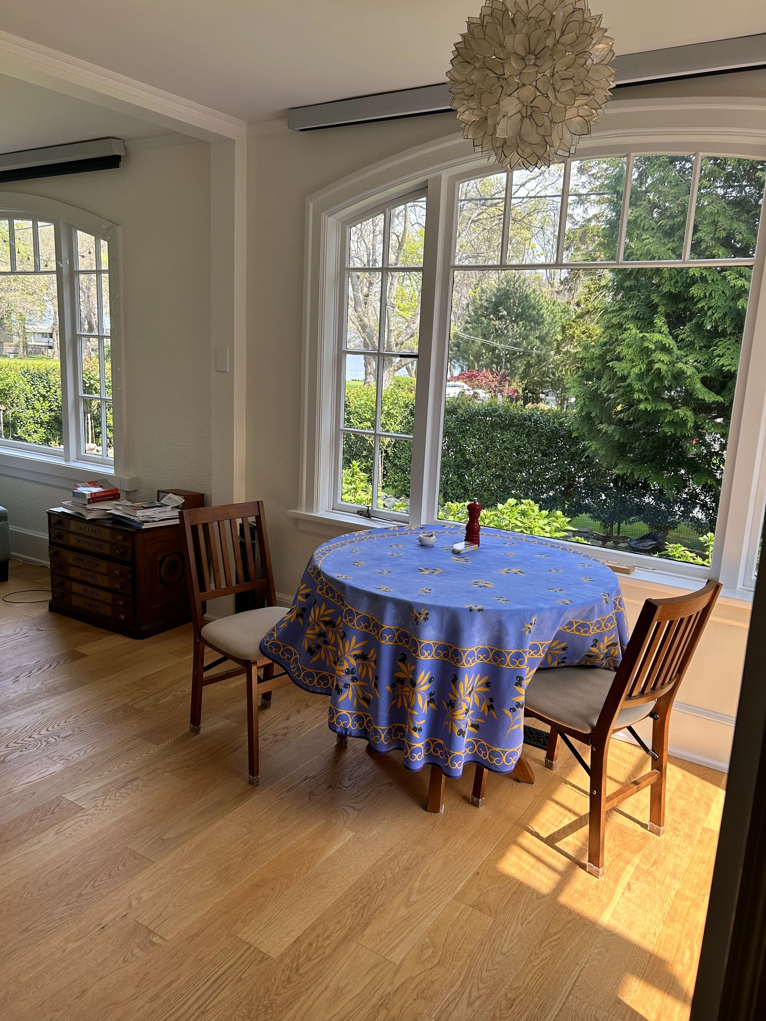 Bright dining area with a round table covered by a blue tablecloth with gold floral patterns, two wooden chairs with beige cushions, and large windows letting in natural light and showing a view of green trees and bushes outside.