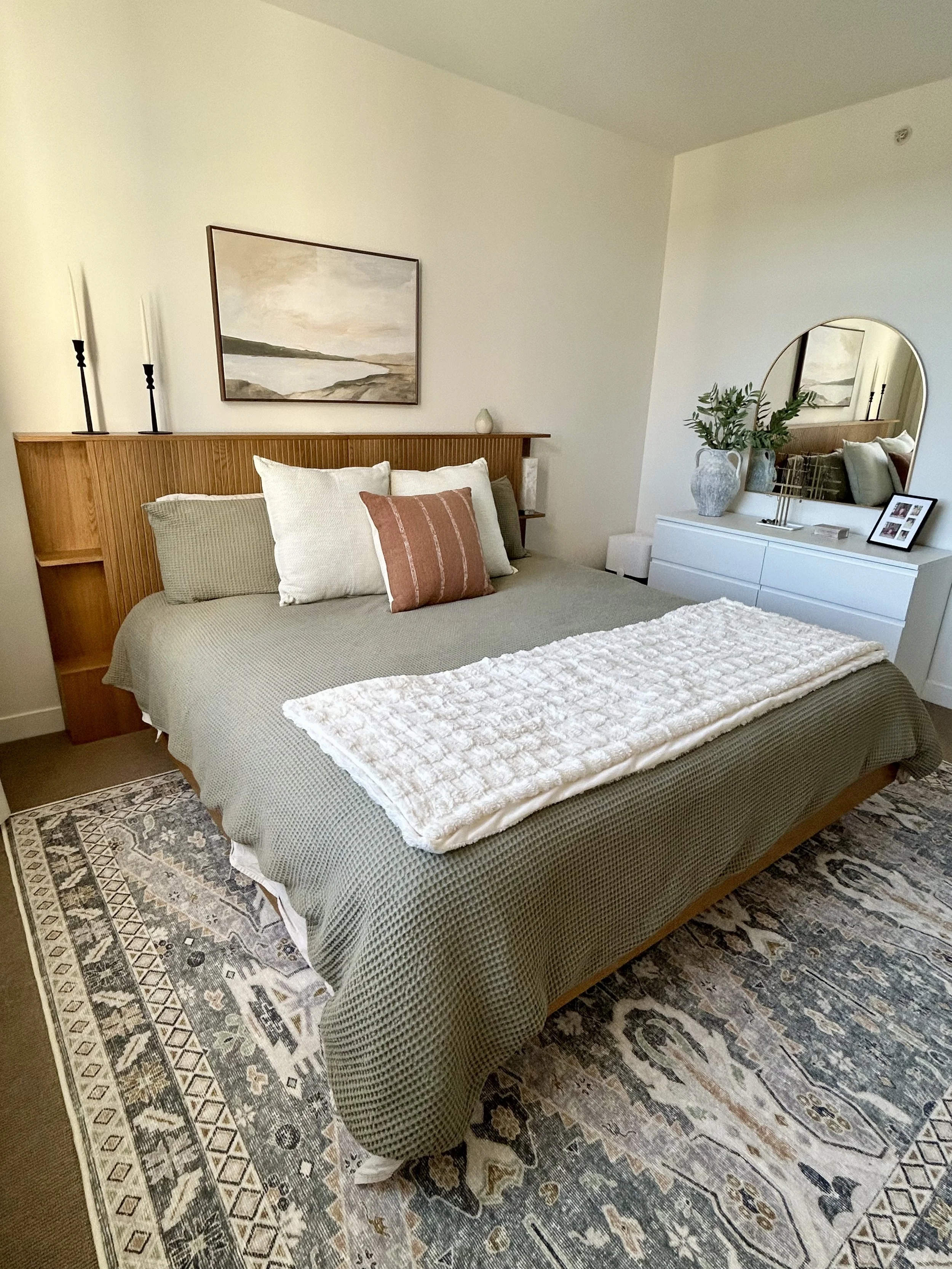 A neatly made bed with green and beige bedding, four pillows, and a textured cream blanket. A wooden headboard with a landscape painting, black candlesticks, and a small white vase. To the right, a white dresser with a large mirror, potted plant, pic