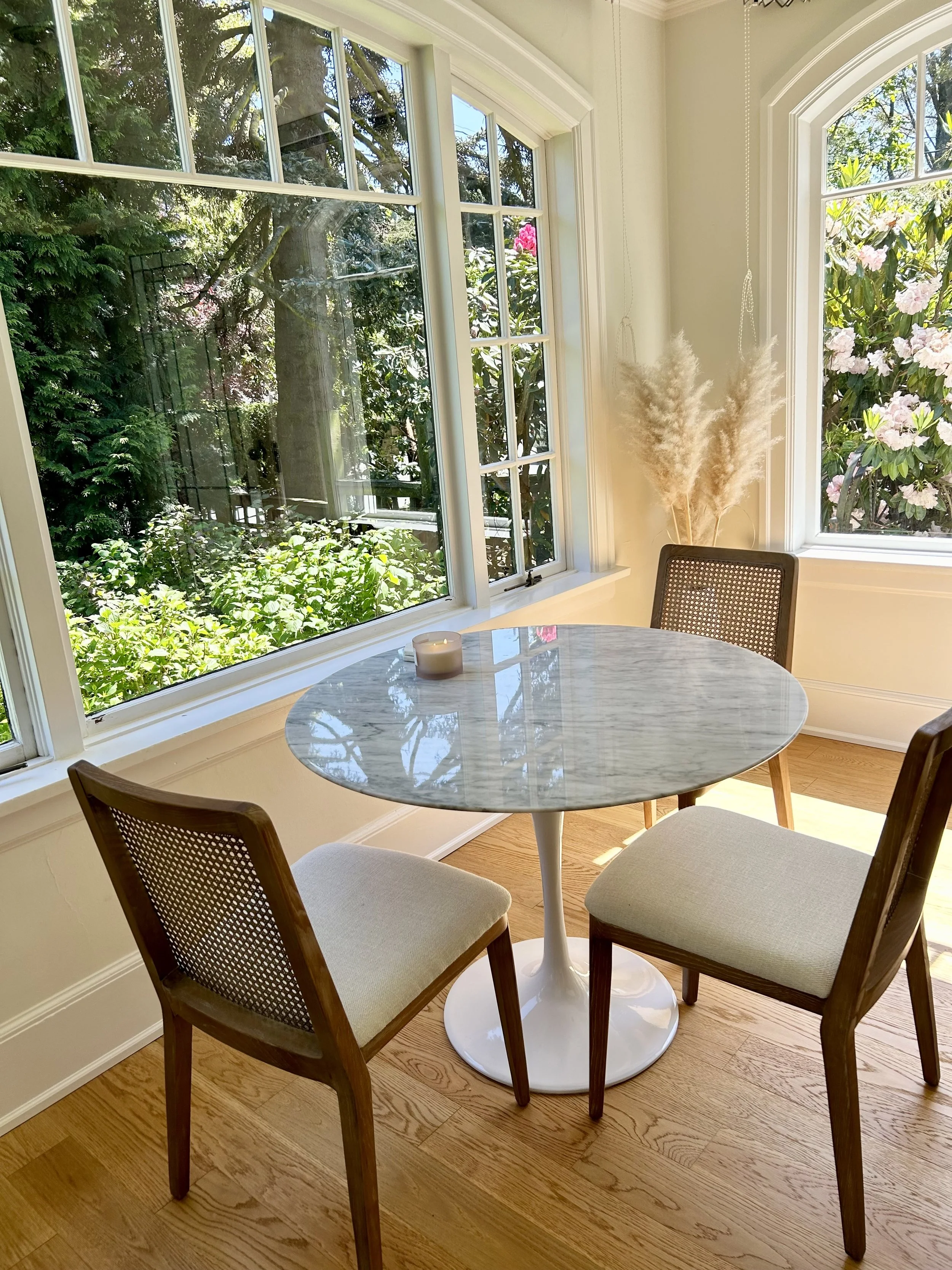 A round marble table with a candle on it, surrounded by two beige upholstered chairs with wooden frames, near large windows showing a garden with green trees and pink flowering bushes.