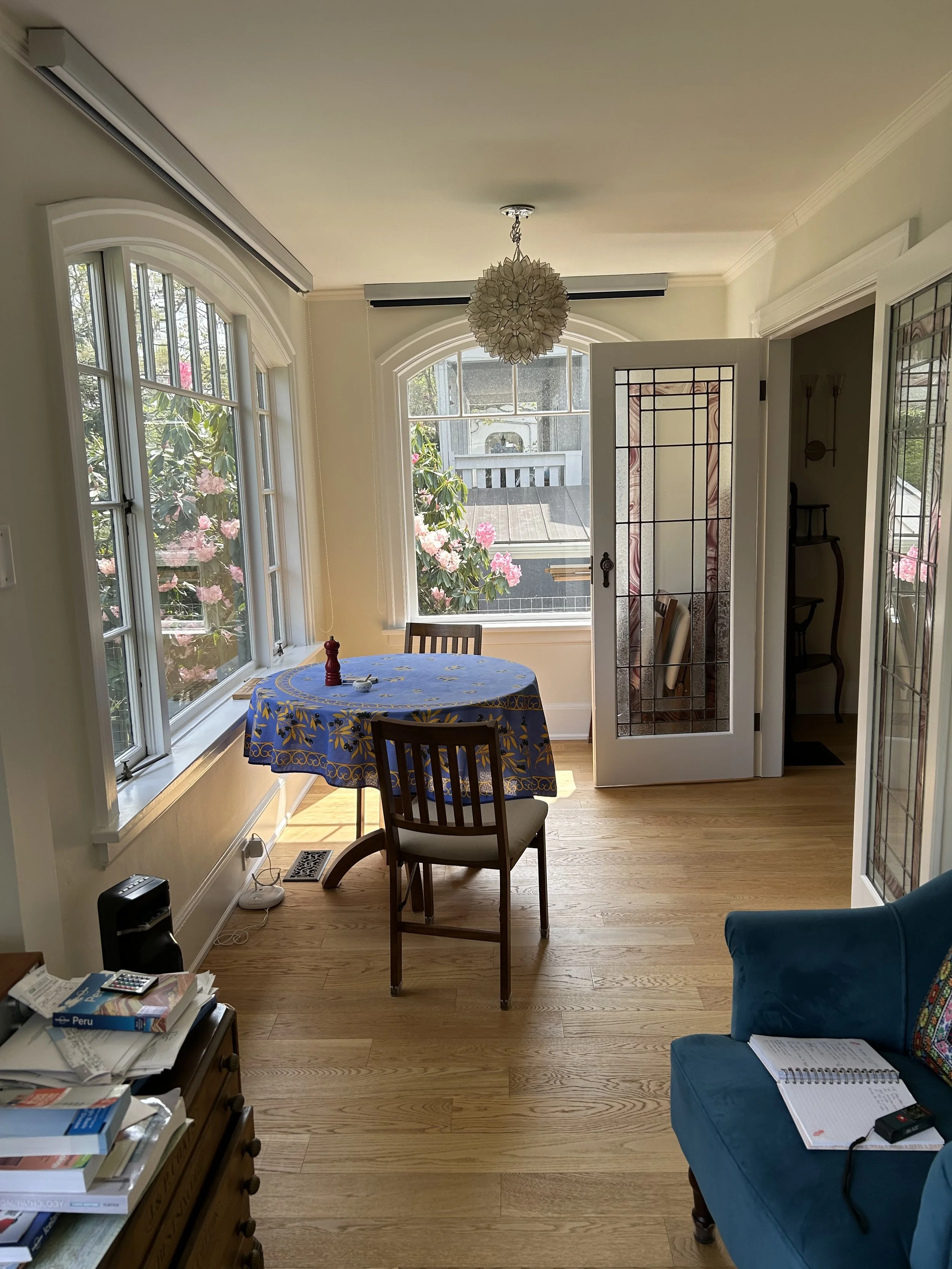 Sunlit dining area with a round table covered by a blue tablecloth, two wooden chairs, large windows with views of pink flowering bushes, and a stained-glass door leading outside.