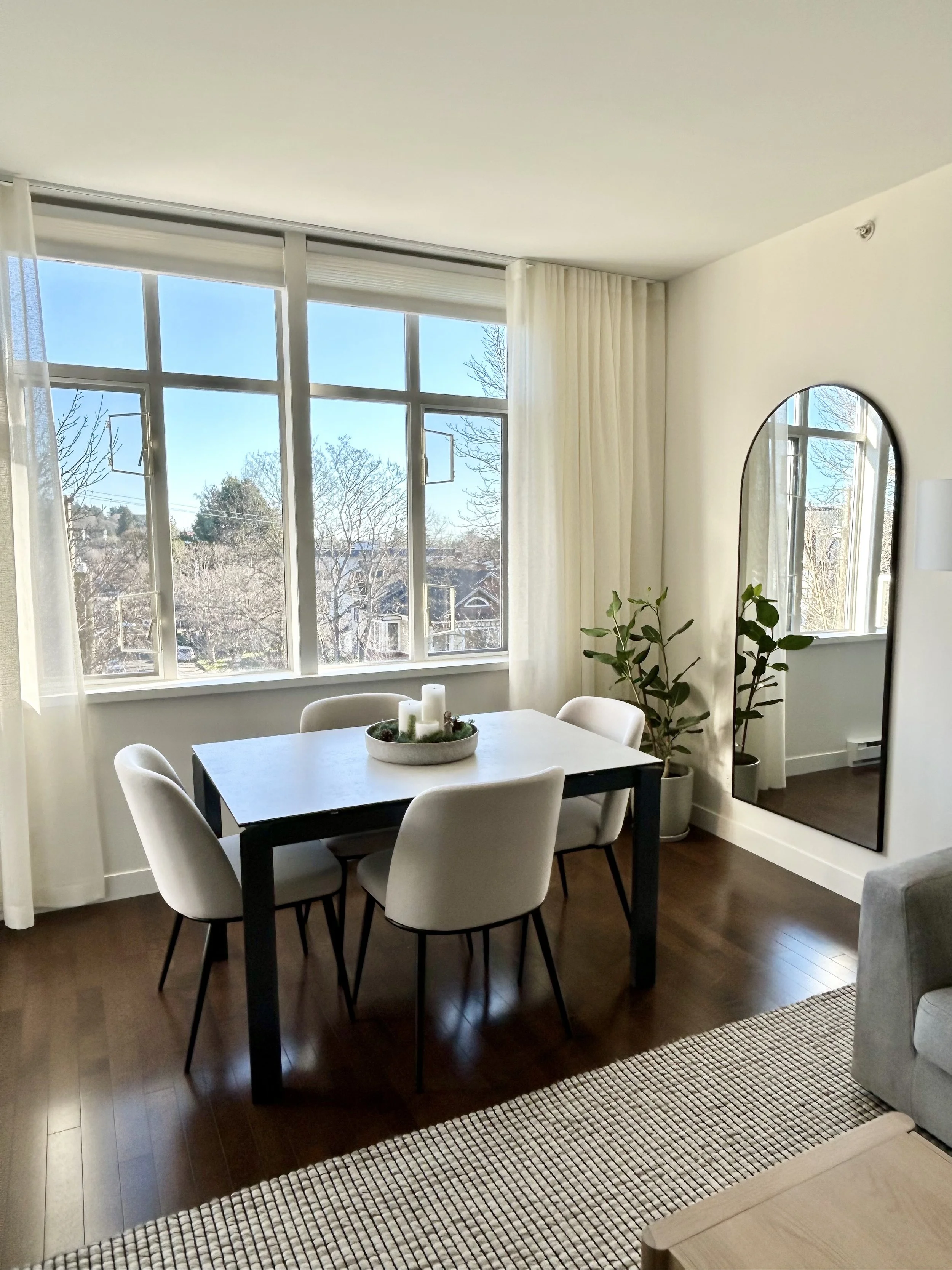 Dining area with a black table, four white chairs, a decorative centerpiece, large window with sheer curtains, potted plants, full-length mirror, and hardwood floors.