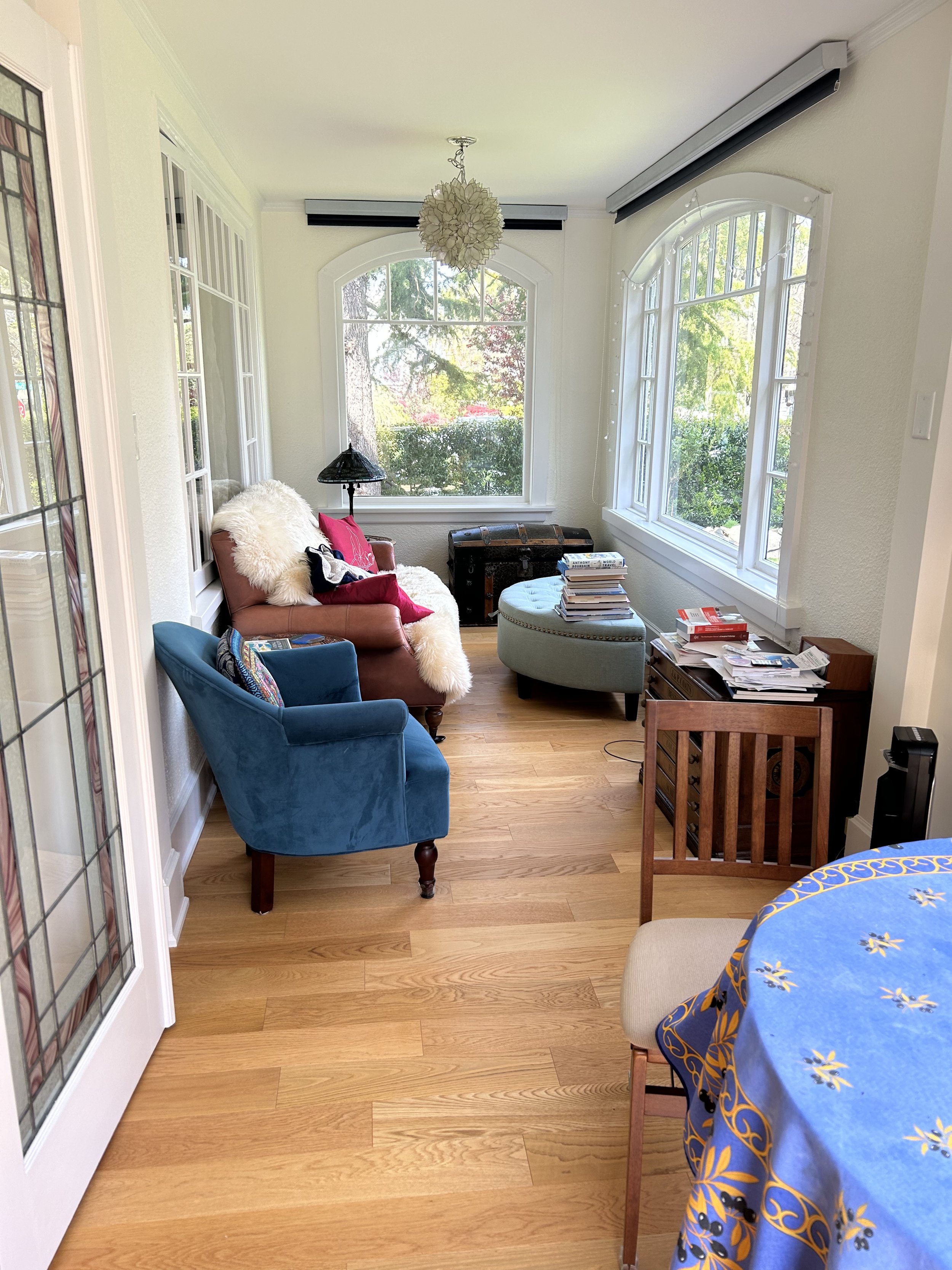 Sunlit porch with vintage furniture, hardwood floors, large windows, and decorative lighting.