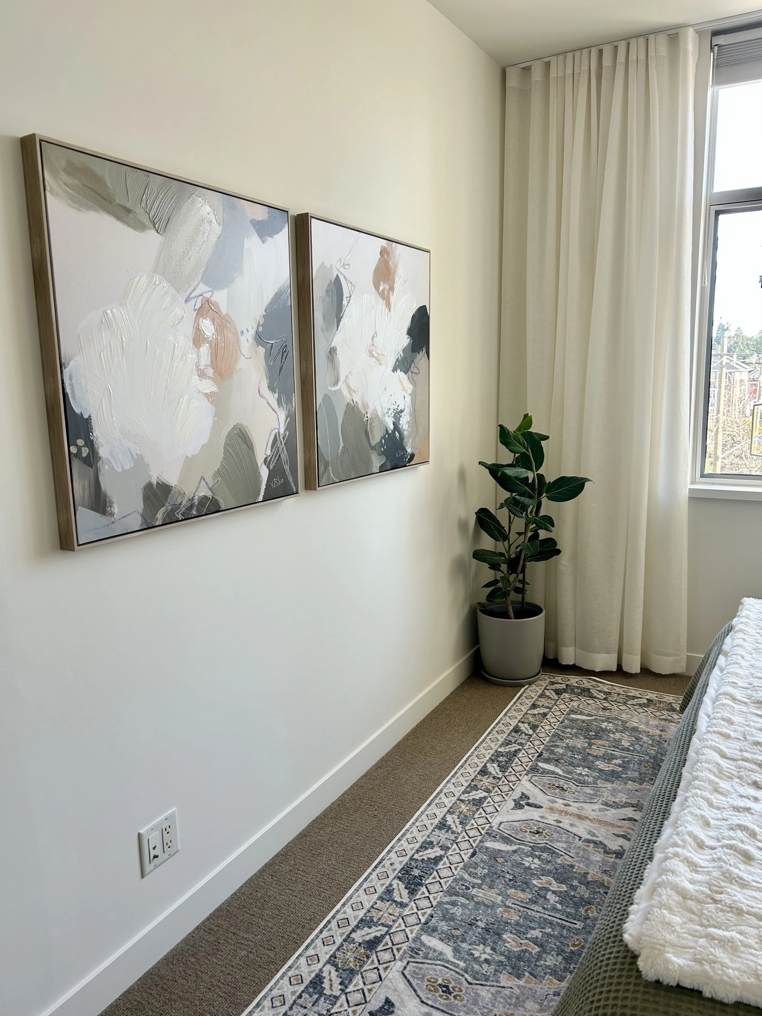 A corner of a bedroom with two abstract paintings on the wall, a potted plant, a window with white curtains, a patterned rug, and part of a bed with a textured white blanket.