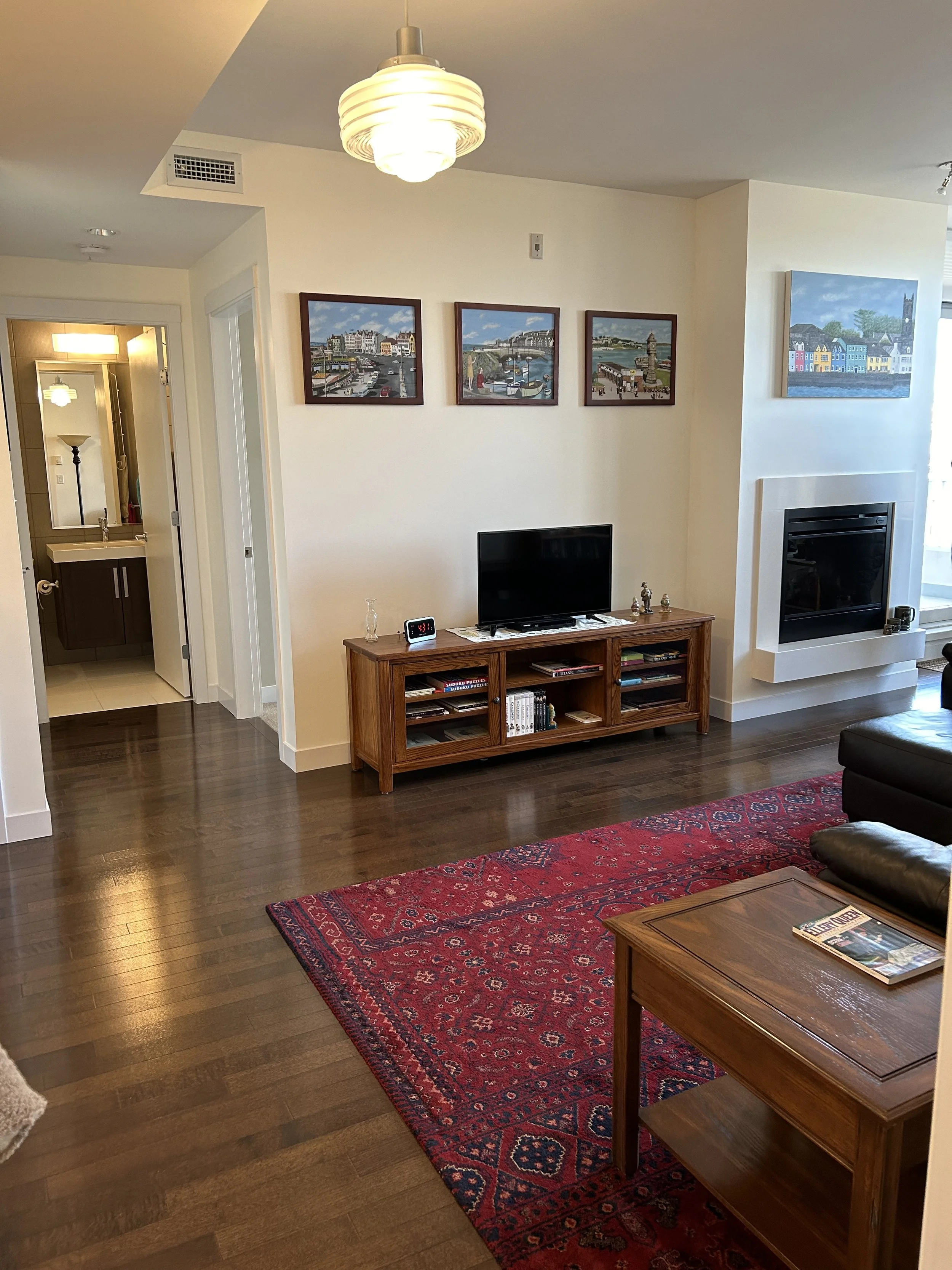 Living room with dark wooden floor, a red patterned rug, a small wooden table with a magazine, a black leather couch, a wooden TV stand with a small flat-screen TV, framed artwork on the wall, a fireplace, and an open doorway leading to a bathroom.