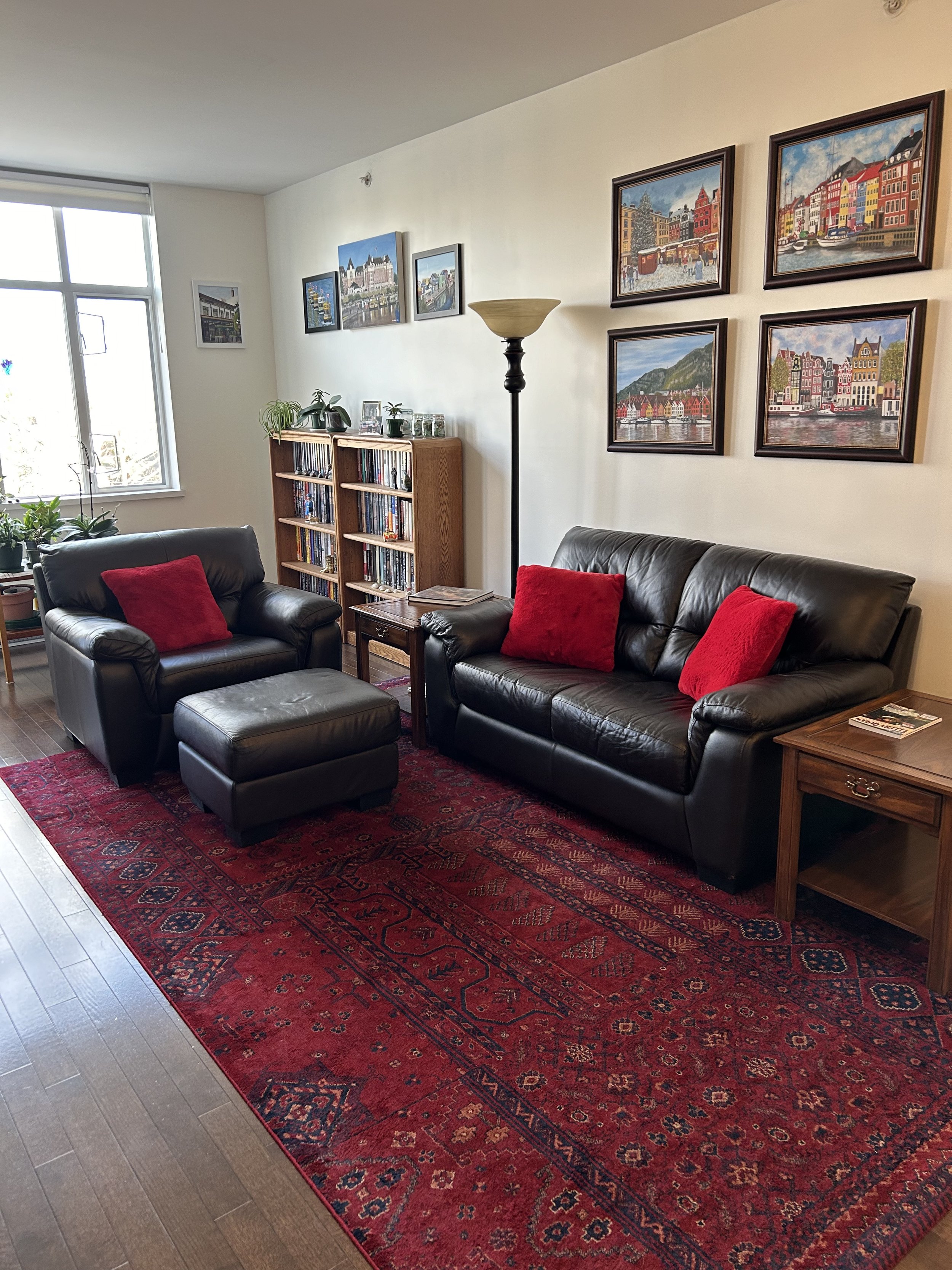 Living room with black leather couch, armchair, and ottoman, all with red pillows. A large red patterned rug covers the hardwood floor. The wall features a series of framed colorful paintings of a cityscape. A wooden bookshelf filled with books and d