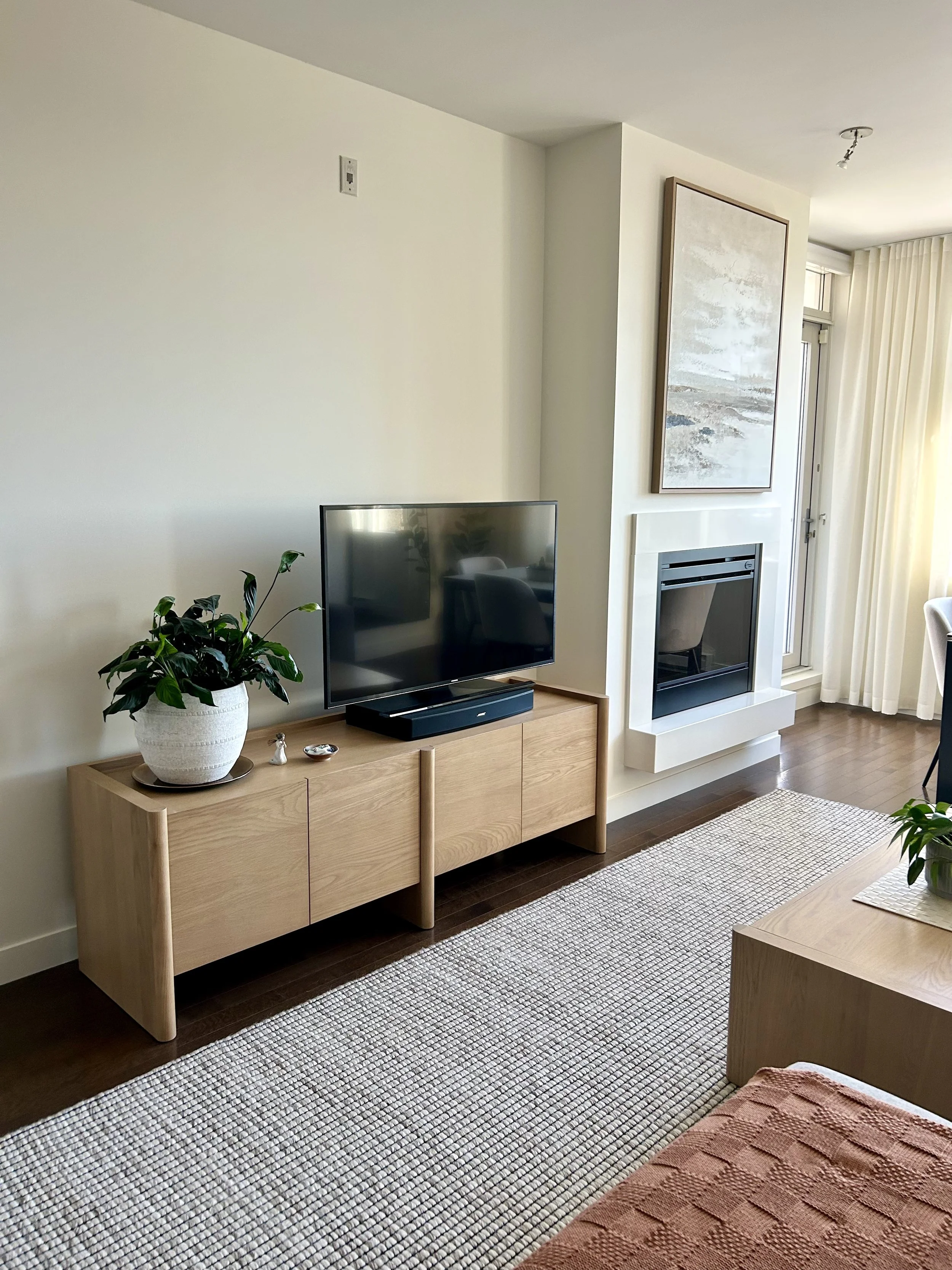 Living room with a wooden TV stand, flat-screen TV, potted plant, fireplace, abstract painting, and curtains.