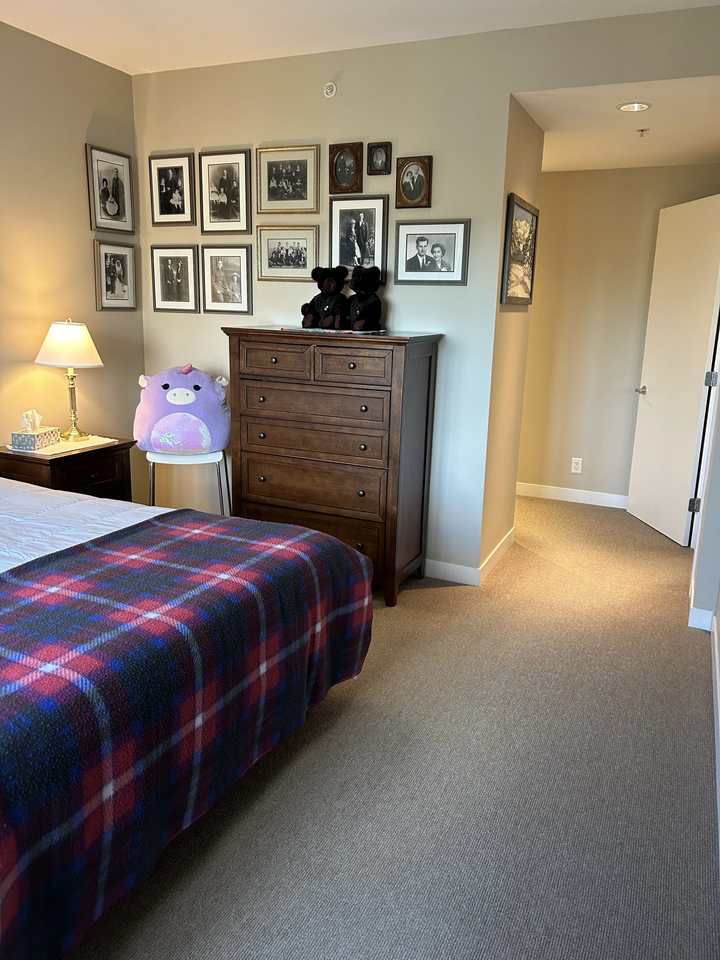 A bedroom with a bed covered in a red and blue plaid blanket, a nightstand with a lamp, a dresser with black teddy bears on top, a plush pillow shaped like a pig on a white chair, and framed photos on the wall.