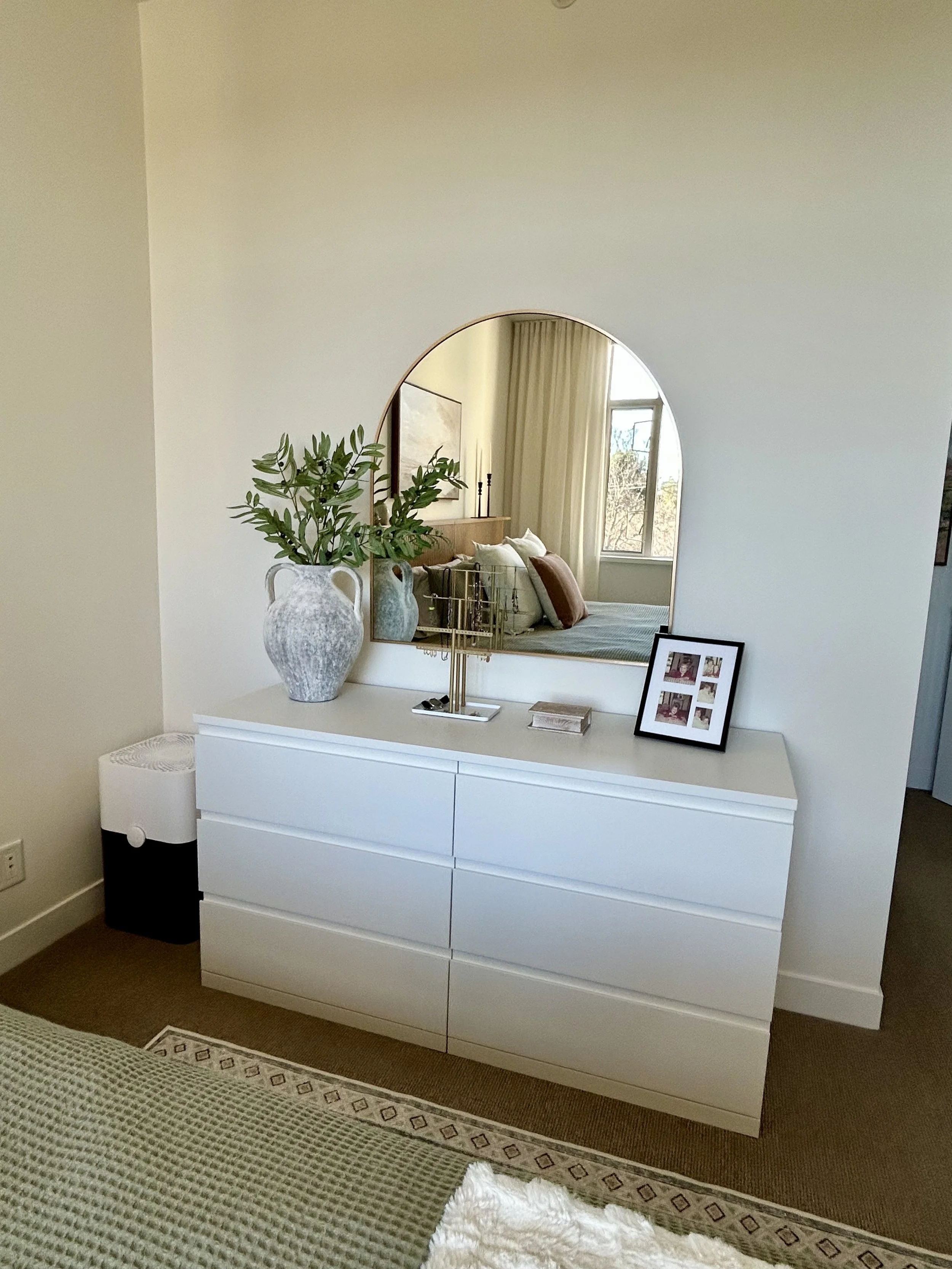 A white dresser with six drawers, topped with a large pottery vase with greenery, a photo frame with multiple pictures, a small gold jewelry stand, and a closed book. A mirror above the dresser reflects part of a bed with pillows and a window with cu