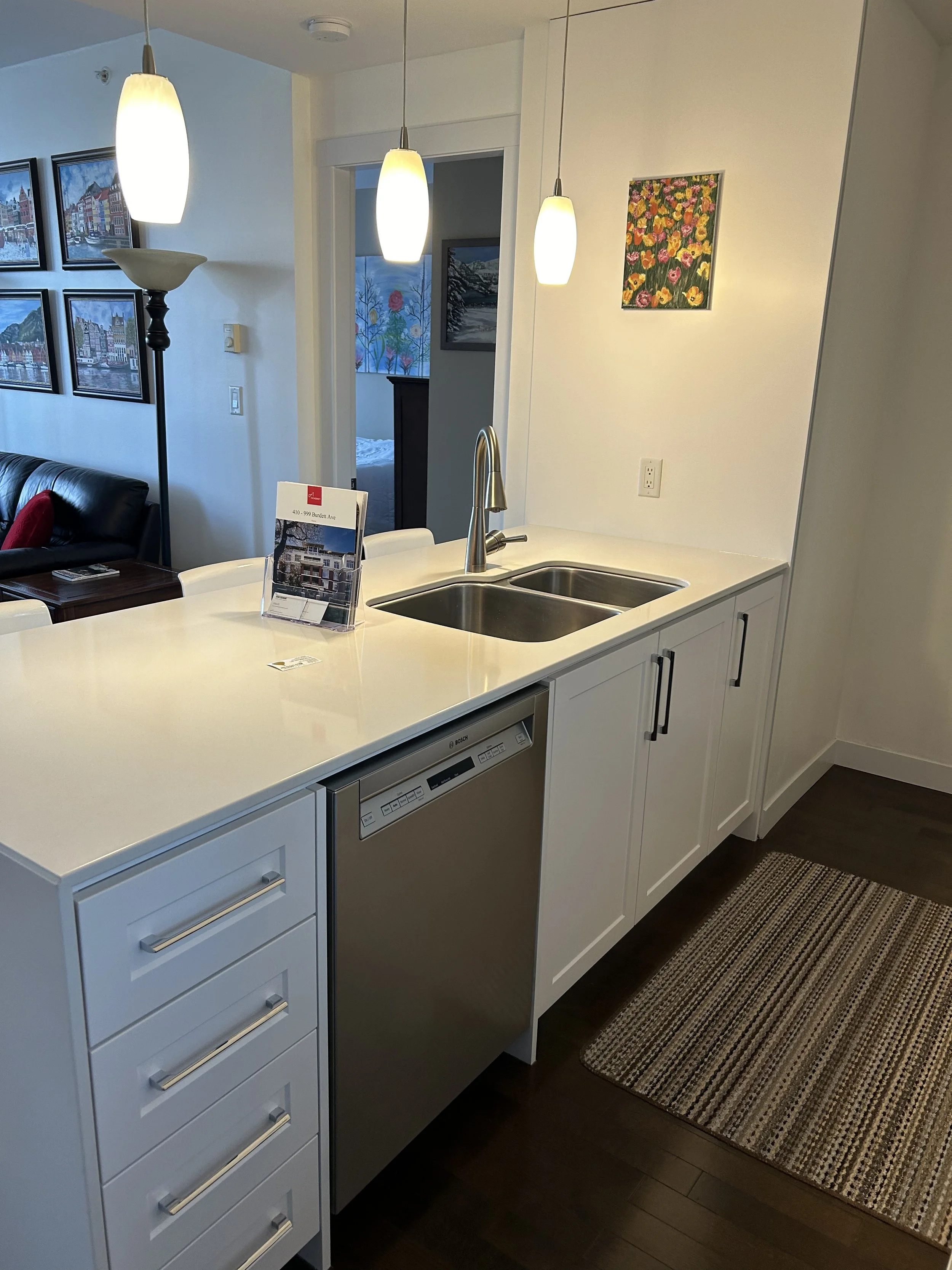 Kitchen with white cabinets, stainless steel dishwasher, double sink, and three pendant lights above the counter. A small decorative picture hangs on the wall.