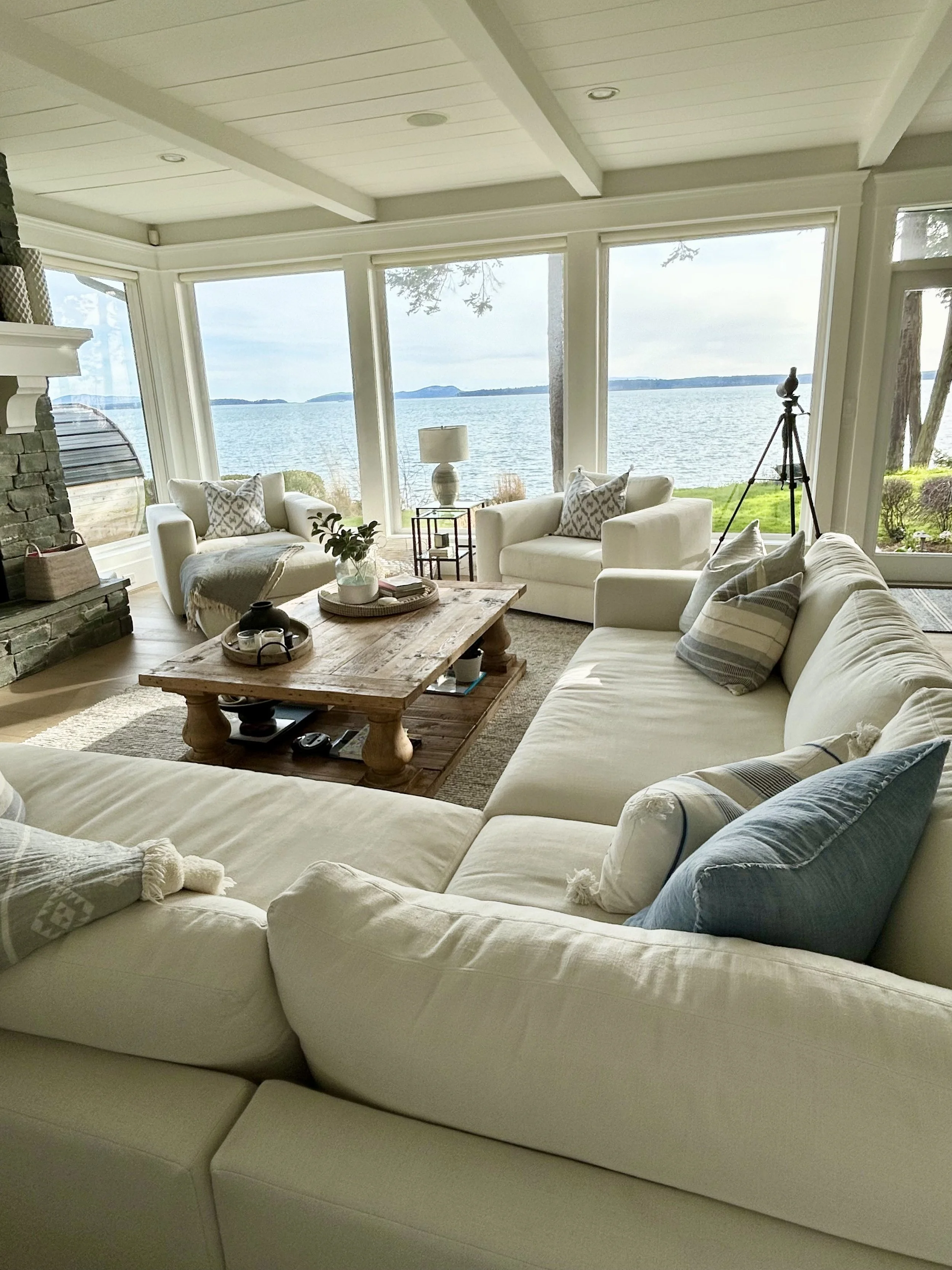 Living room with large windows overlooking a body of water, white furniture including a sectional sofa with striped pillows, coffee table with decorative items, armchairs, a lamp, and a telescope.