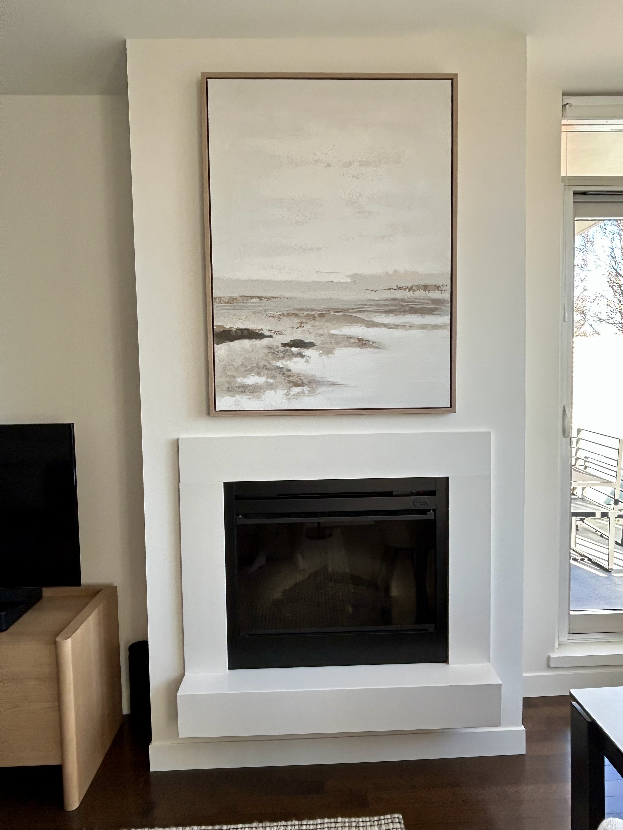 A modern fireplace with a white mantel and a black glass front, located on a white wall. Above the fireplace hangs a large abstract landscape painting in neutral tones. To the right, there is a glass door leading to an outdoor balcony with a metal ra
