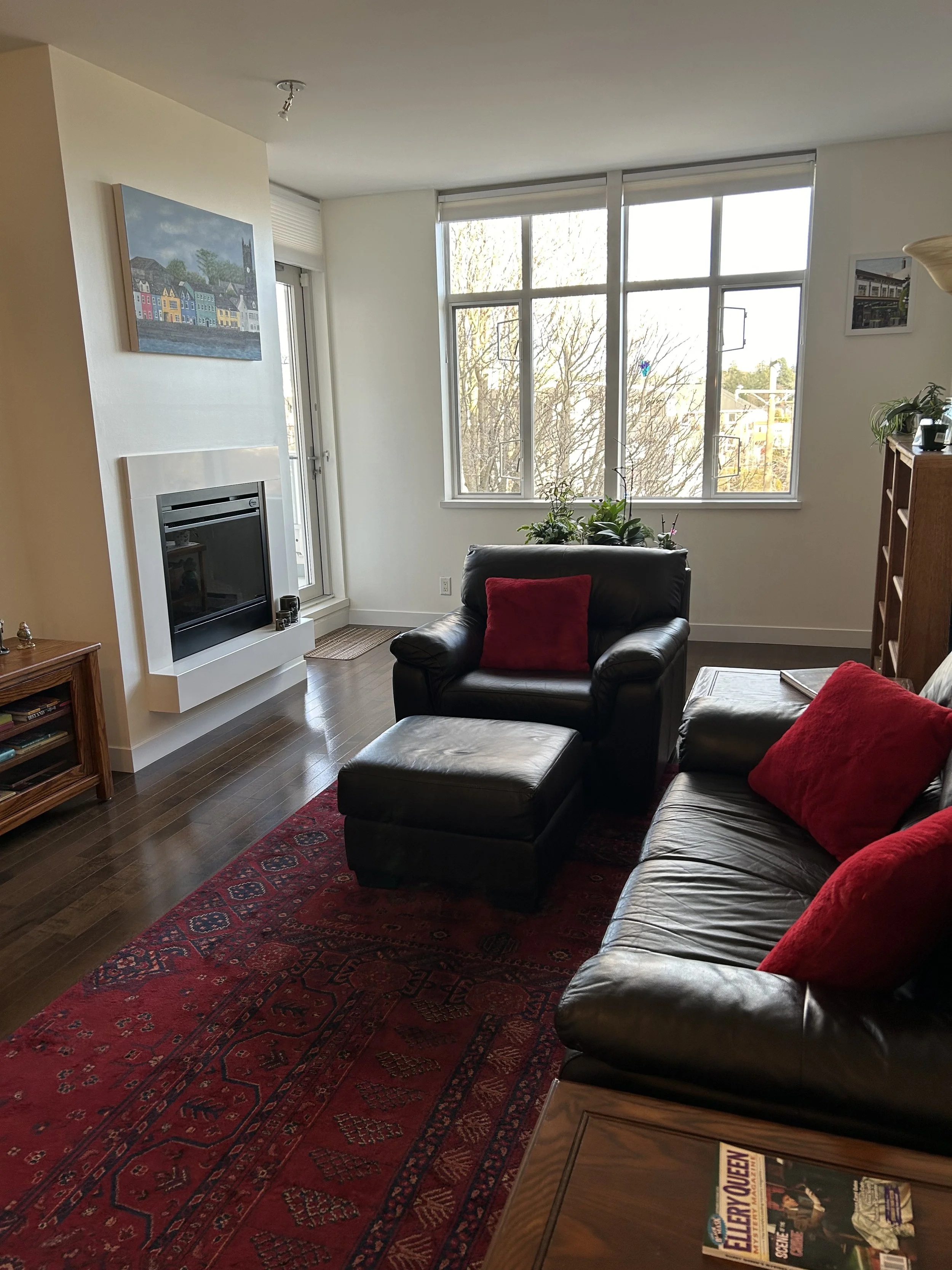 Living room with black leather sofa and matching armchair, red pillows, wooden furniture, large window with daylight, potted plants, red patterned rug, fireplace, books, and artwork on the walls.
