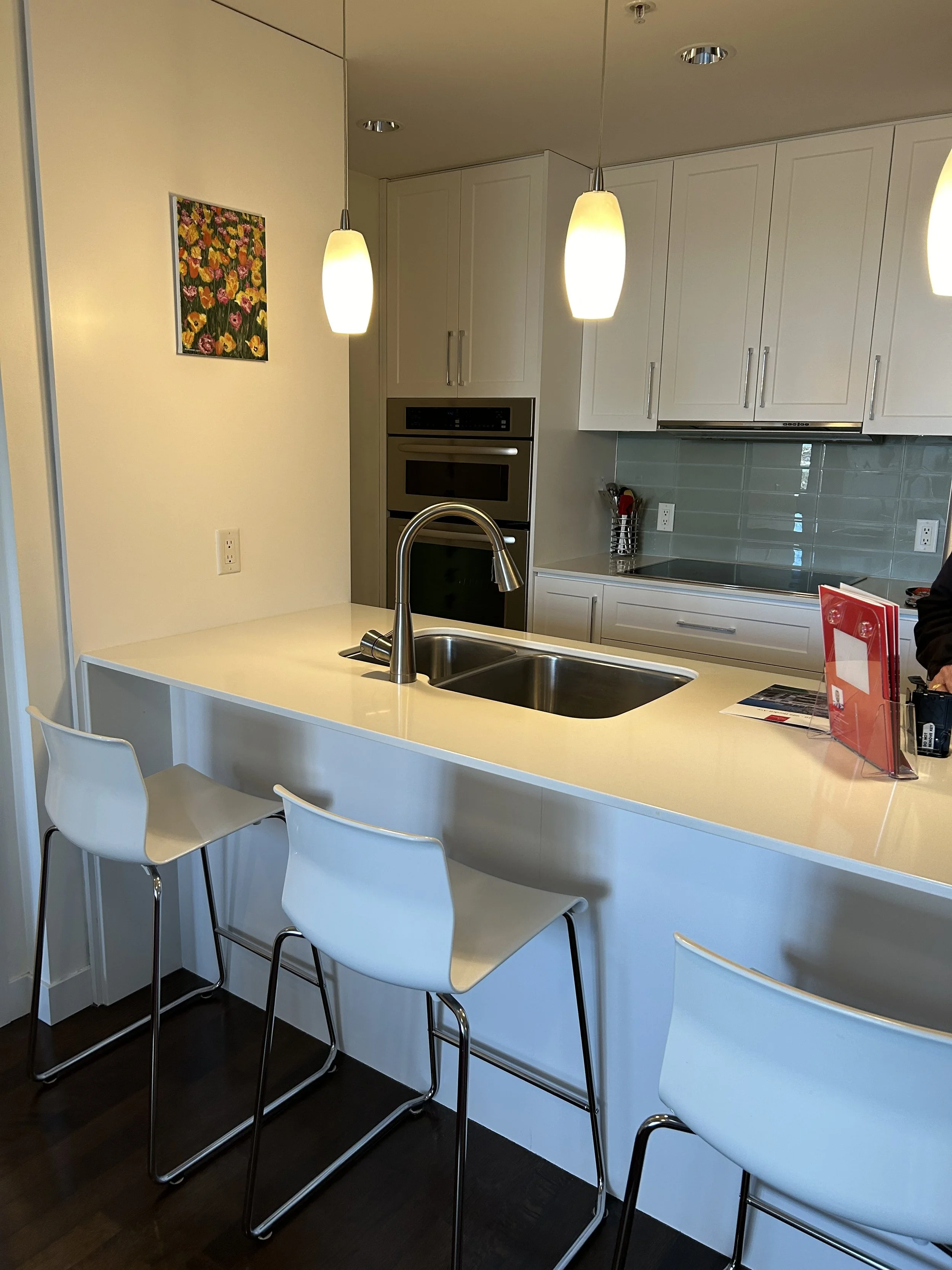 Modern kitchen with white cabinets, stainless steel appliances, a double sink, pendant lighting, and a breakfast bar with three white chairs.