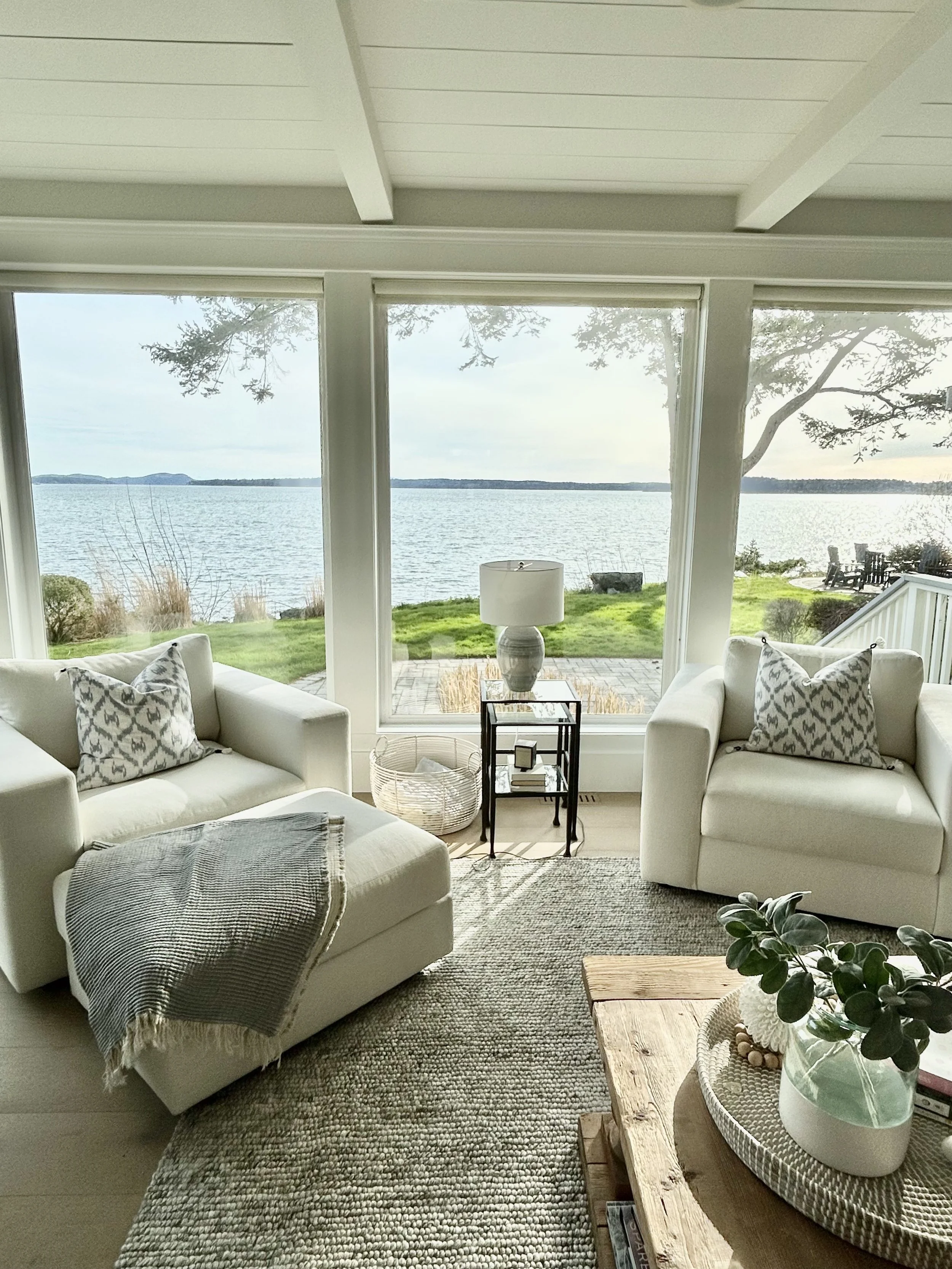 Living room with large windows overlooking a lake, featuring white sofas, patterned pillows, a wooden coffee table with a plant, and a side table with a lamp, with a grassy yard and trees outside.