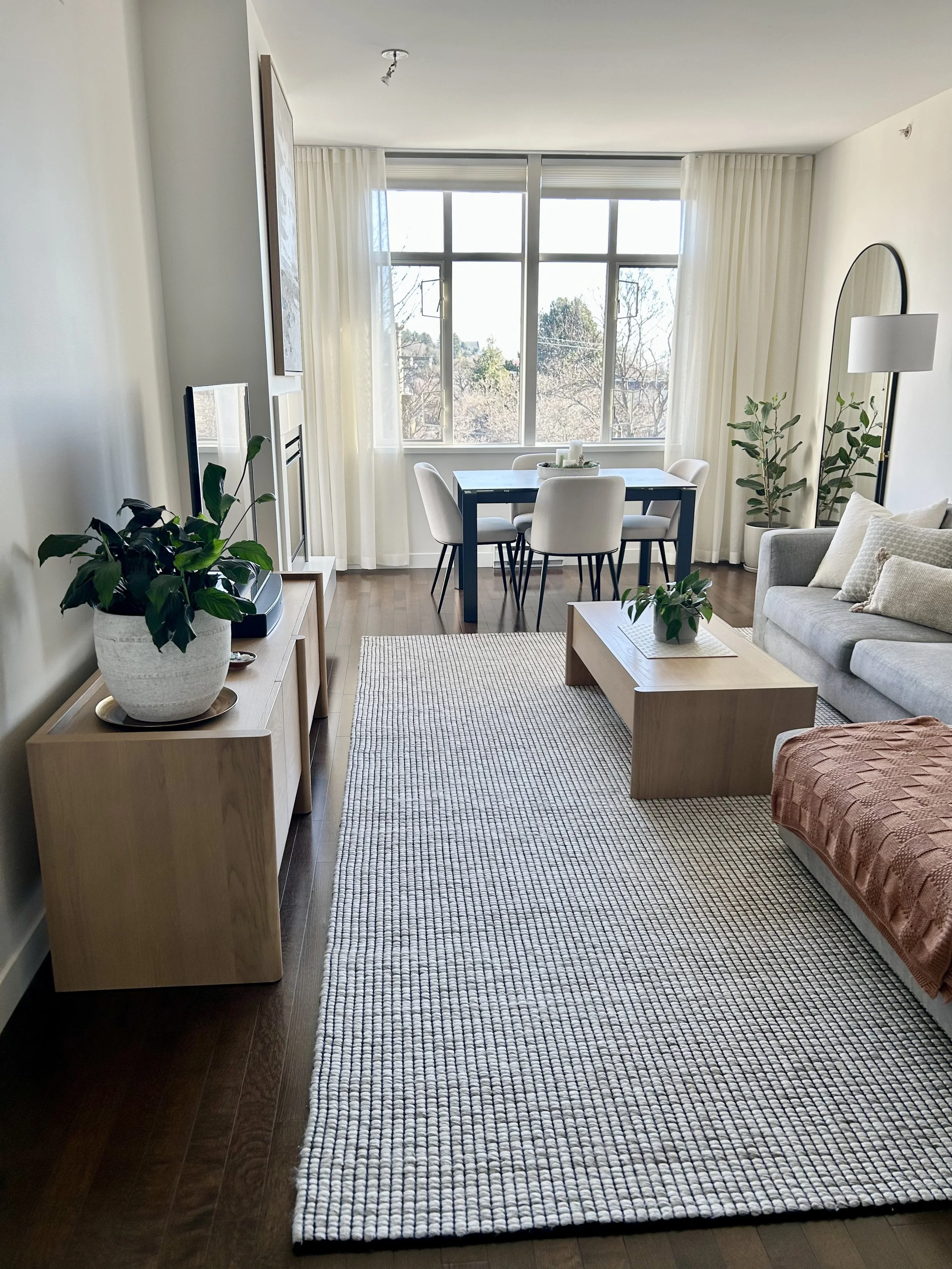 Bright living room with a large window, gray sofa with pillows, wooden coffee table, plants, a TV on a wooden stand, dining table with white chairs, and a small rug on wooden floor.