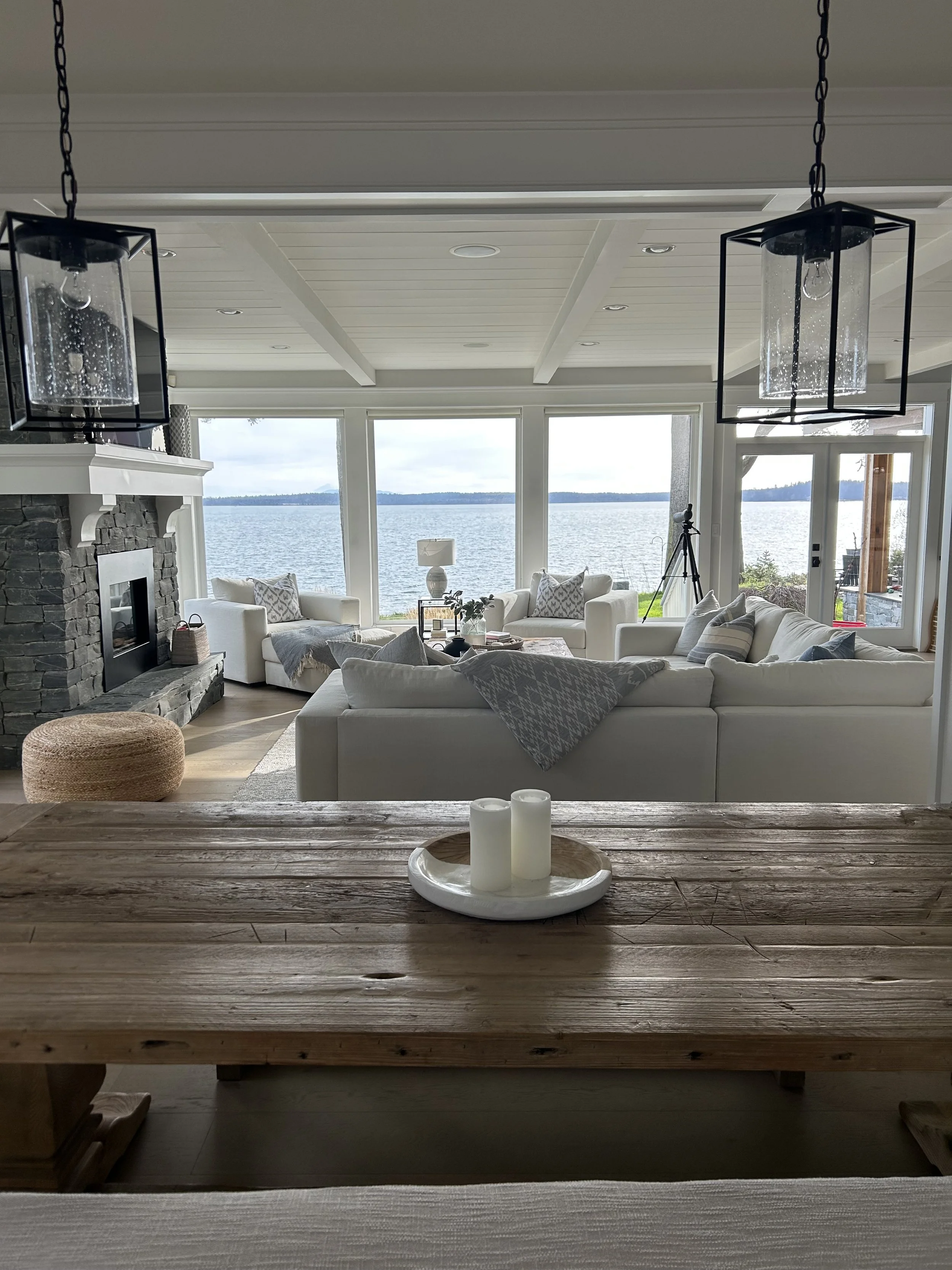 Living room with white sofas, a stone fireplace, large windows overlooking a body of water, and nautical decor.