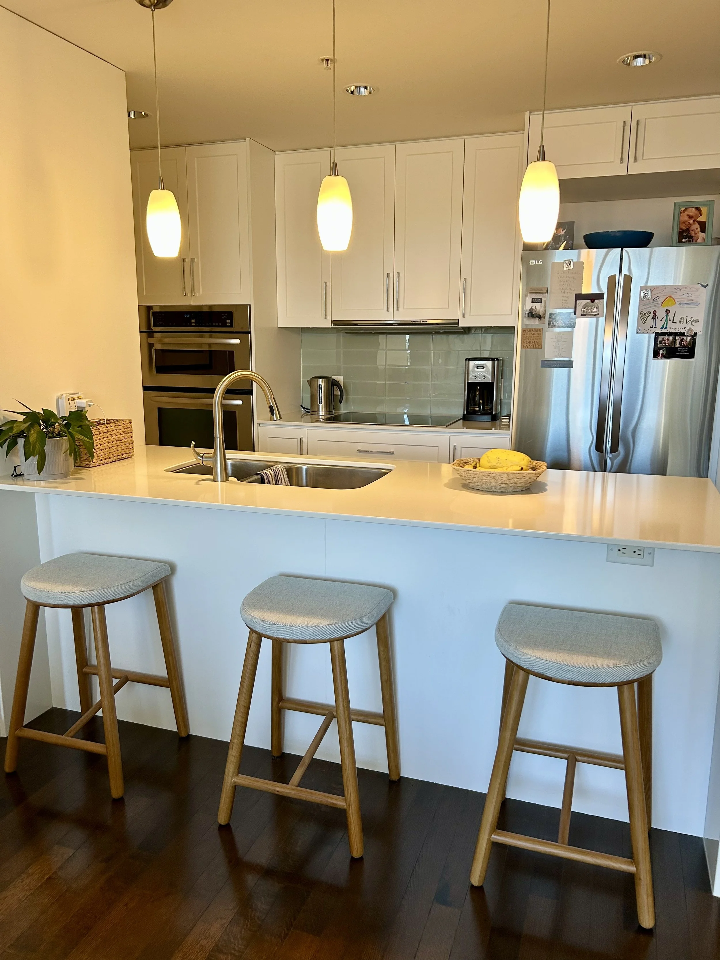 Modern kitchen with white cabinets, a green tile backsplash, stainless steel appliances, a large stainless steel refrigerator with photos and notes on it, a breakfast bar with three wooden stools, pendant lighting, and a dark wood floor.