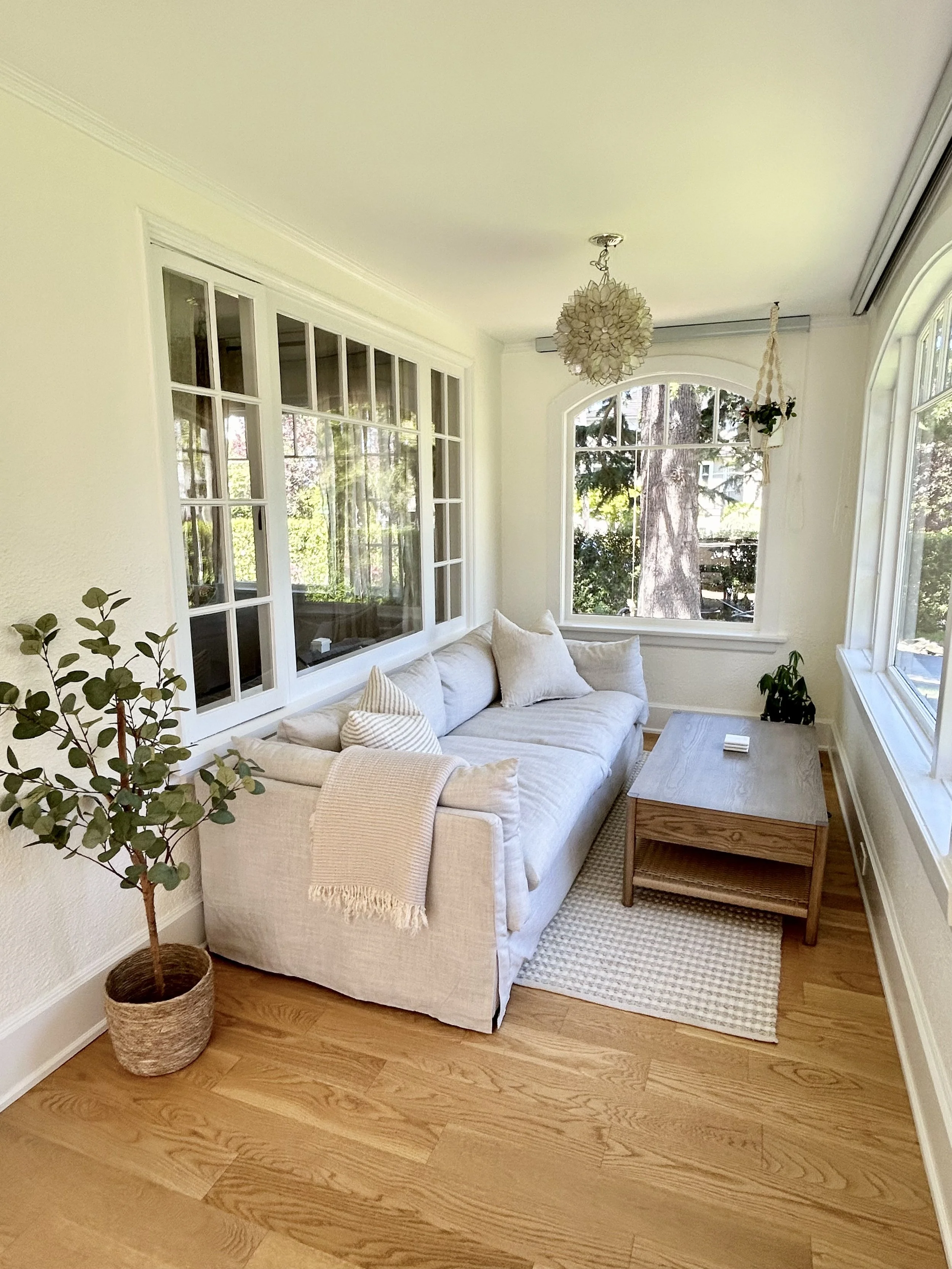 Bright living room with large front windows, white sofa with cushions and blanket, wooden side table, potted plant, hanging light fixture, and view of trees outside.
