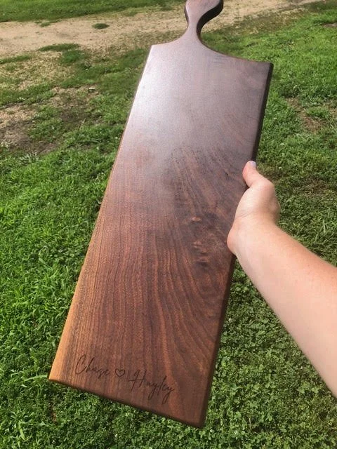 Long dark hardwood charcuterie board with a decorative handle and personalized engraving, held outdoors against a grassy background.