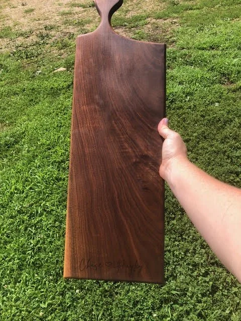 Handcrafted long wooden serving board with rich dark grain, a rounded handle, and personalized engraving near the bottom.