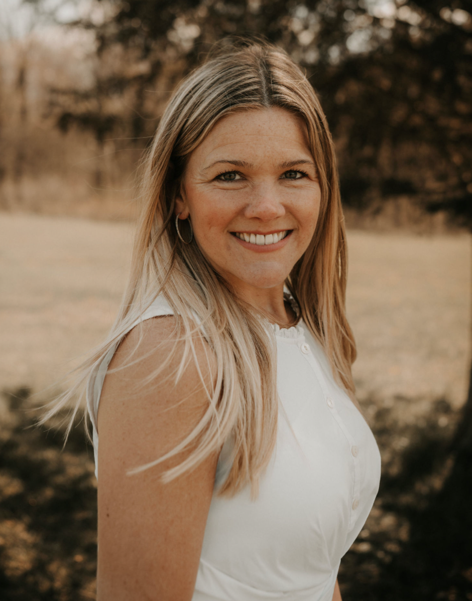 A smiling woman with blonde hair, wearing a white sleeveless blouse, standing outdoors in a park during fall with trees in the background.