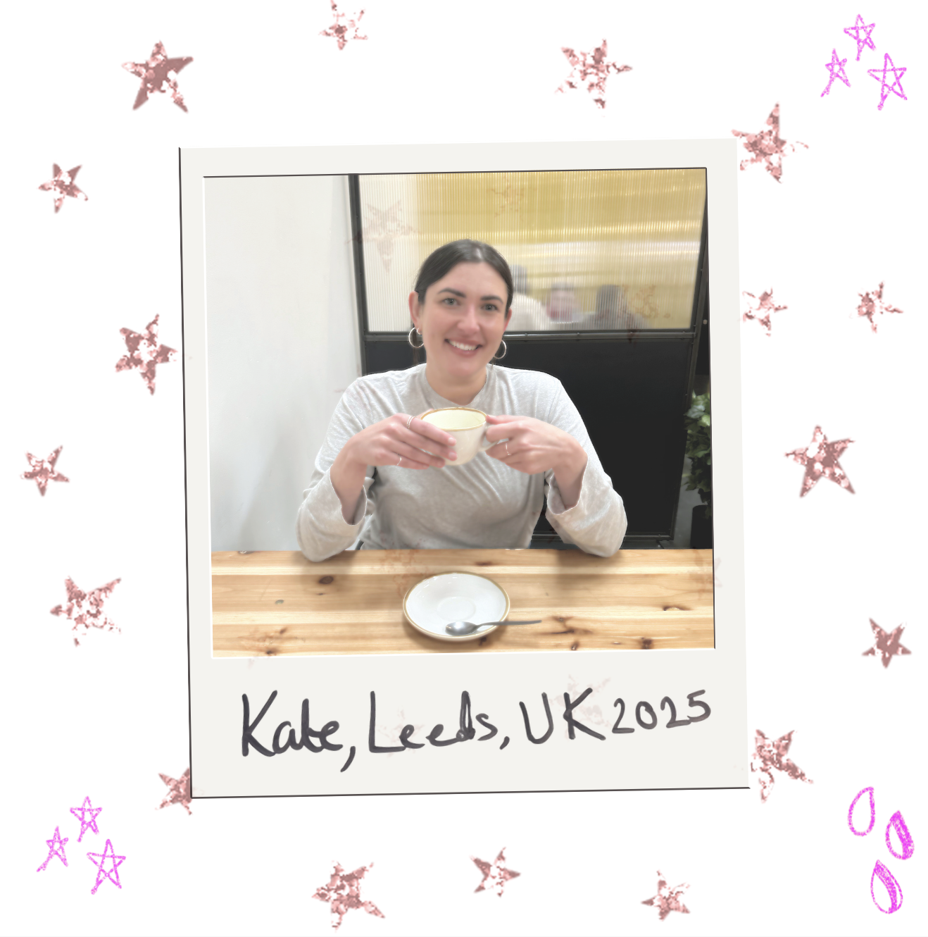Kate, freelance UGC creator and social media strategist from Leeds, UK, 2025, smiling at a wooden table while holding a white cup. Dark-haired with hoop earrings, surrounded by pink and red stars, with a window in the background.