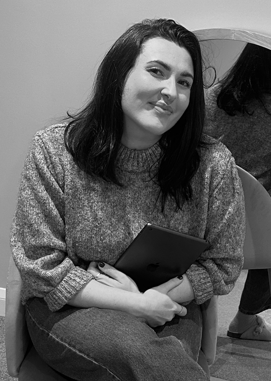 A young woman with dark hair wearing a sweater, sitting on a chair, holding a tablet, and smiling at the camera.