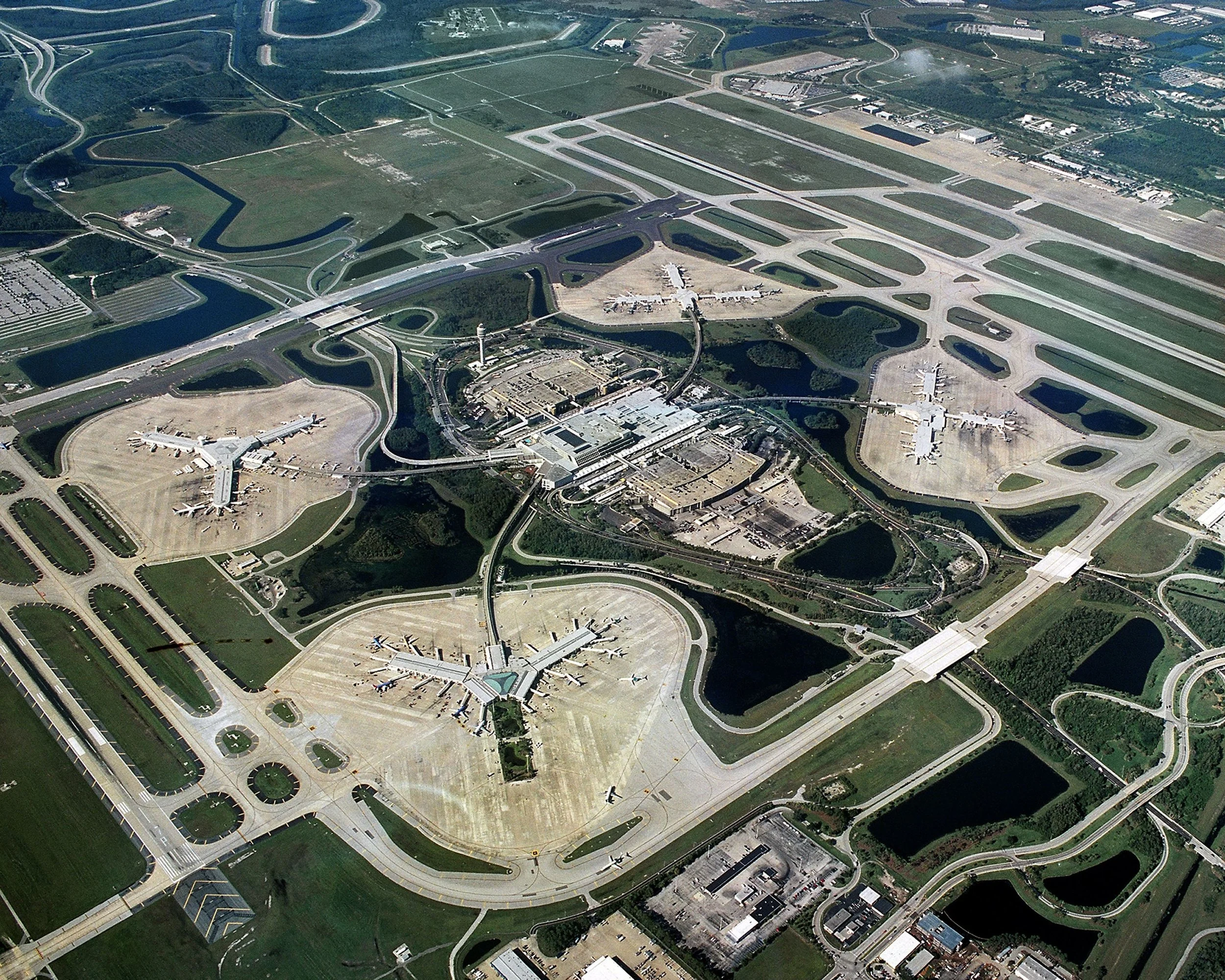 Aerial view of an airport with multiple runways, taxiways, and terminal buildings surrounded by green parks, lakes, and roads.