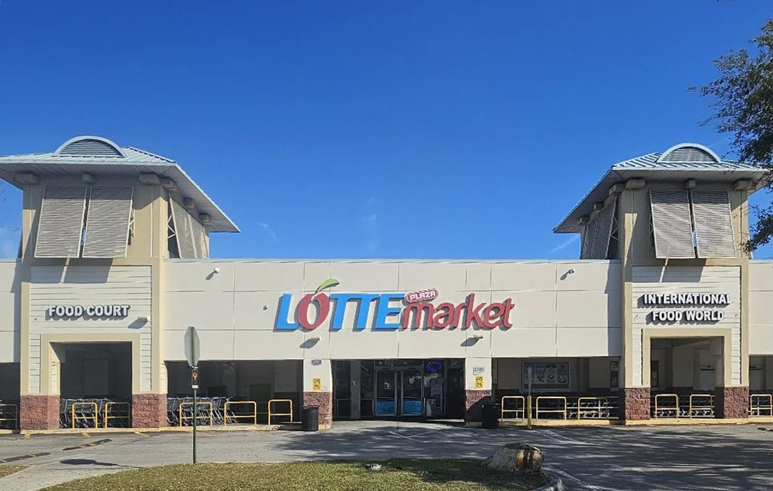 Front view of a Lotte Mart hypermarket with a blue sky, featuring the store's sign, entrance doors, and surrounding pavement.