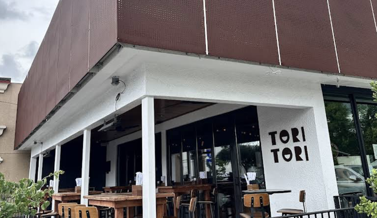 Exterior view of a restaurant named Tori Tori with outdoor seating, wooden tables and chairs, and a white wall with the restaurant's name.