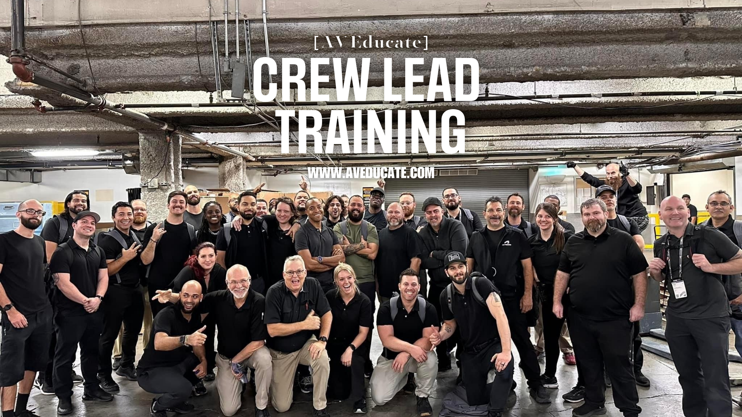 A large group of people, diverse in age, gender, and ethnicity, gathered in an industrial setting. They are smiling and posing for a group photo, some making peace signs, with a sign overhead that reads 'AV Educate Crew Lead Training' and a website URL 'www.aveducate.com'.