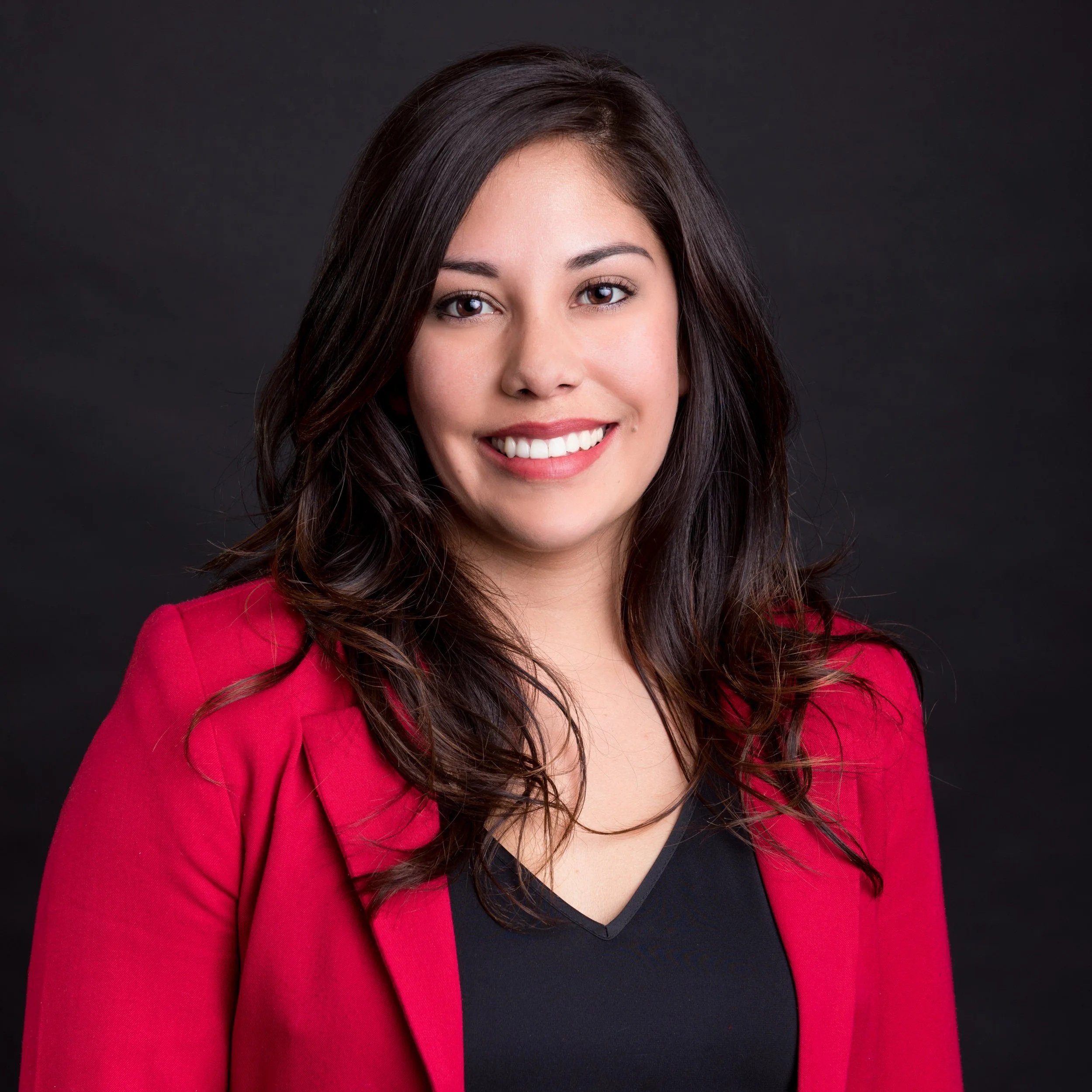 Headshot of a young woman with long dark brown hair, smiling, wearing a black top and red blazer, against a dark gray background.