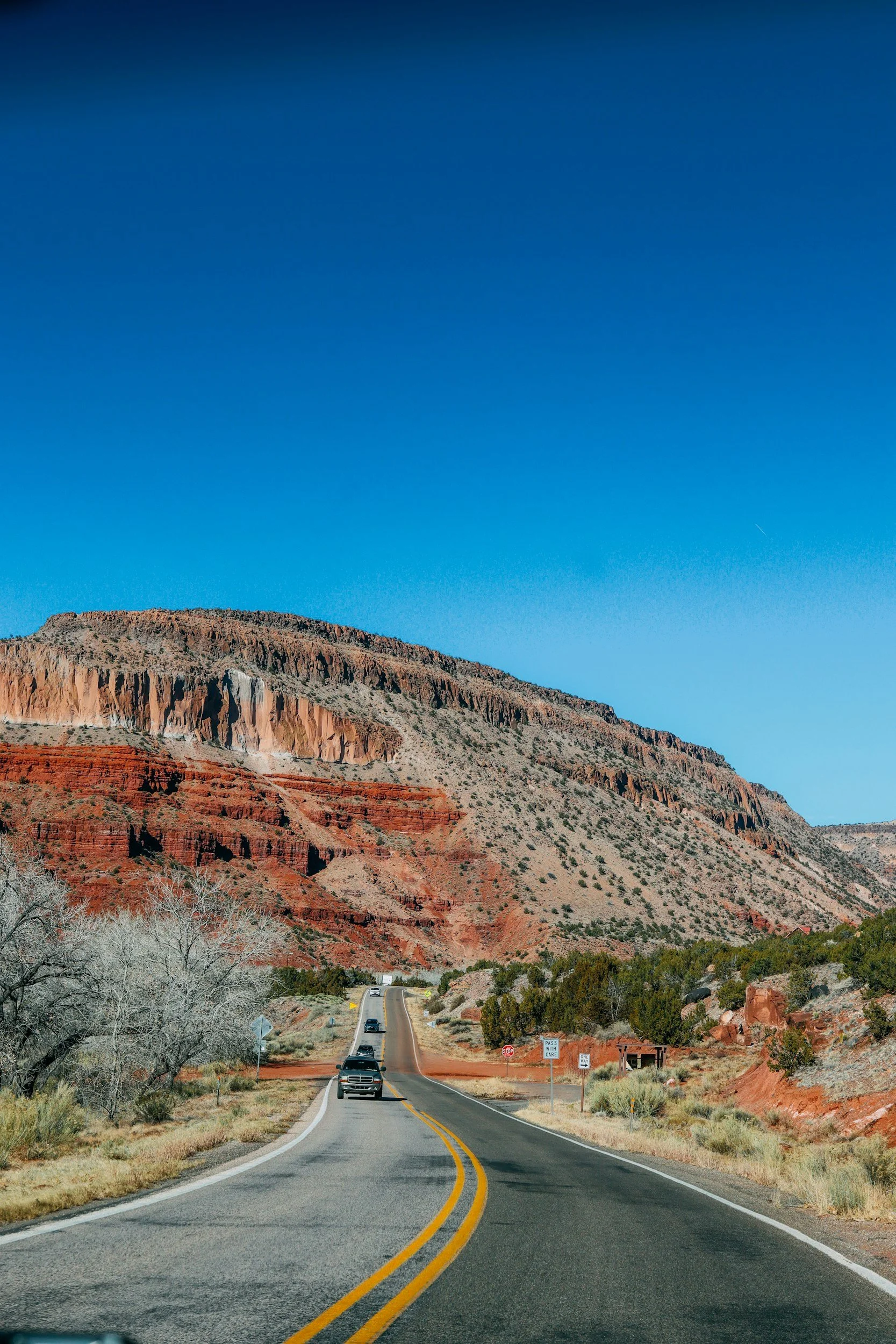 A winding two-lane road with yellow lines, traveling through a desert landscape with red and orange layered cliffs, sparse vegetation, and a clear blue sky.