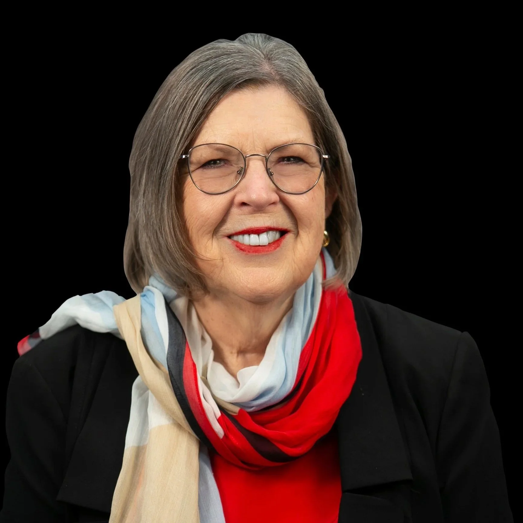 A smiling older woman with gray hair and glasses, wearing a red blouse, a black jacket, and a multi-colored scarf, against a black background.