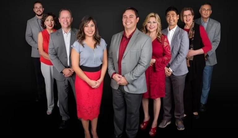 Group of nine diverse professionals posing together in front of a black background, smiling, dressed in business casual attire.