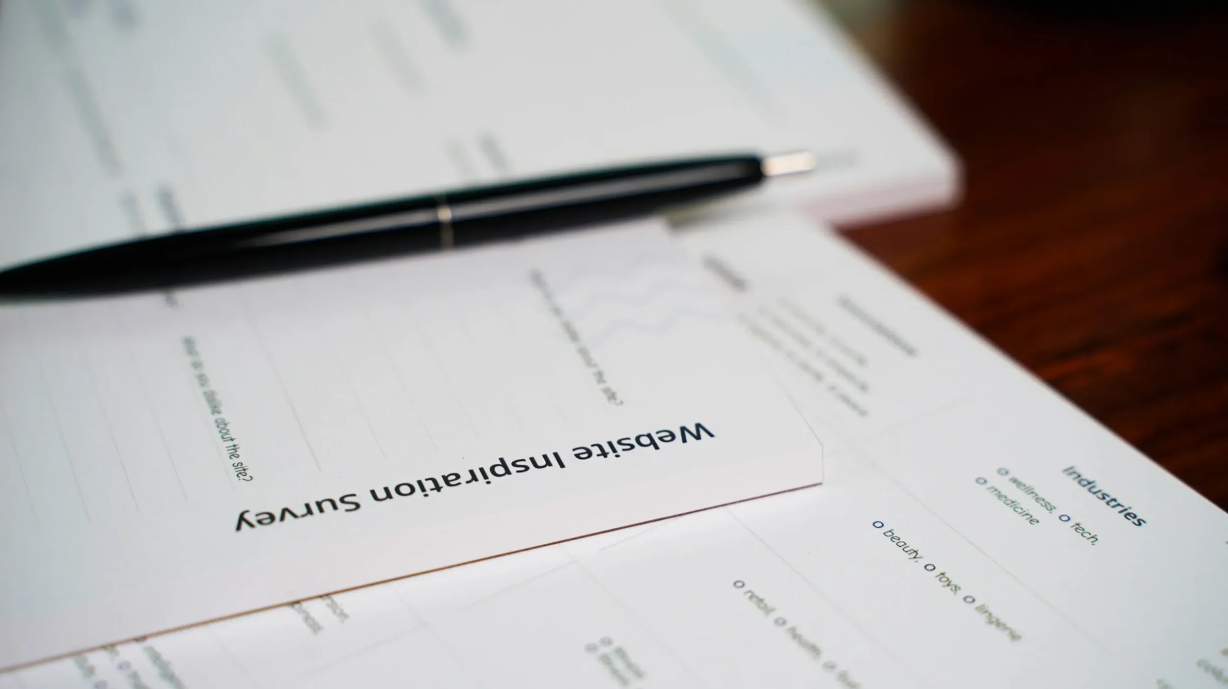 A report titled 'Website Inspiration Survey' on a sheet of paper with a black pen placed on top, and a second sheet with a list of industries and categories including wellness, tech, medicine, beauty, toys, and travel.