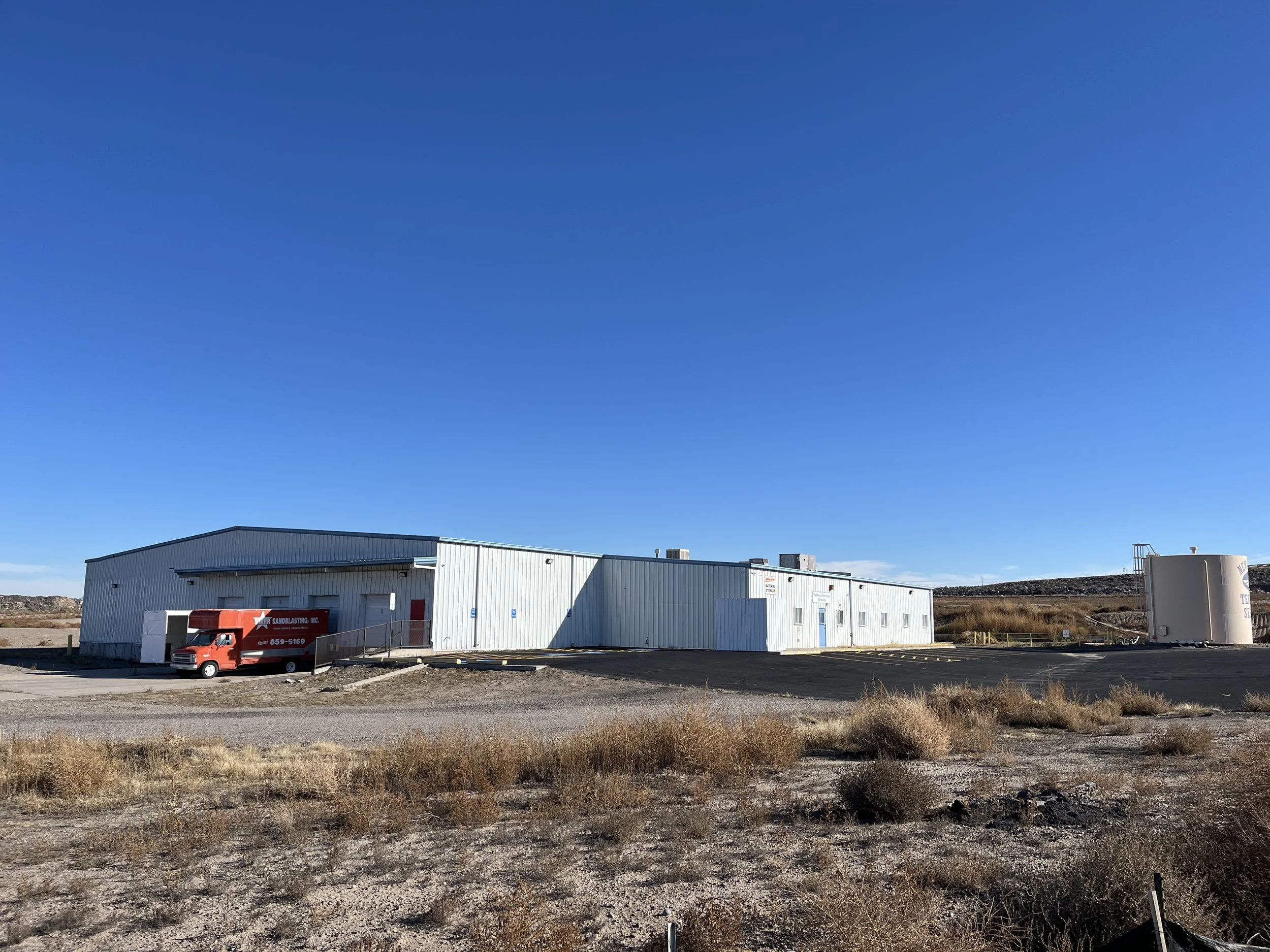 A large industrial building with metal siding, situated in a desert landscape with dry shrubs and clear blue sky, featuring a red van parked near an entrance.