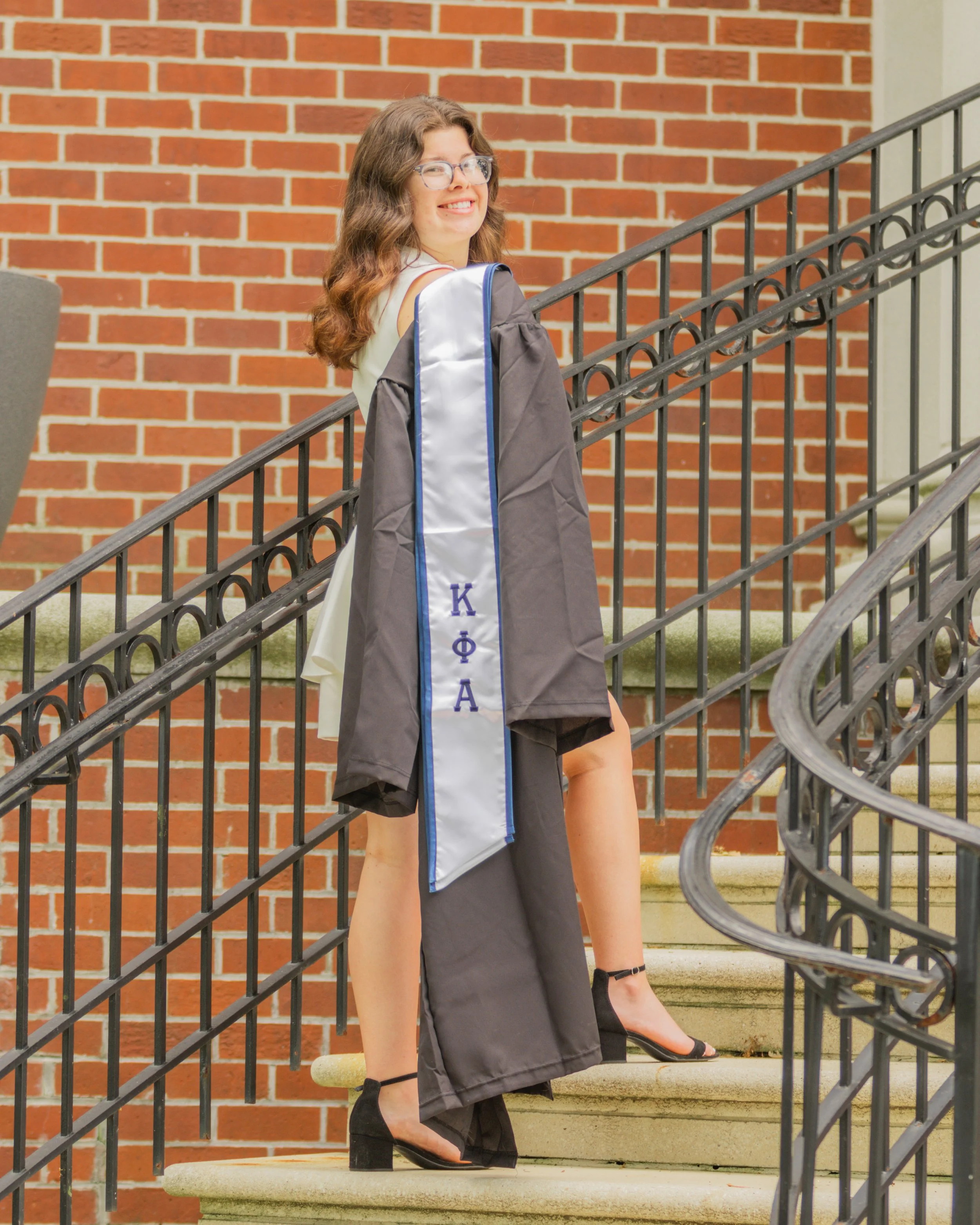 A young woman in graduation cap and gown standing on a staircase, smiling at the camera. She is wearing glasses, black heels, and has a sash with Greek letters.