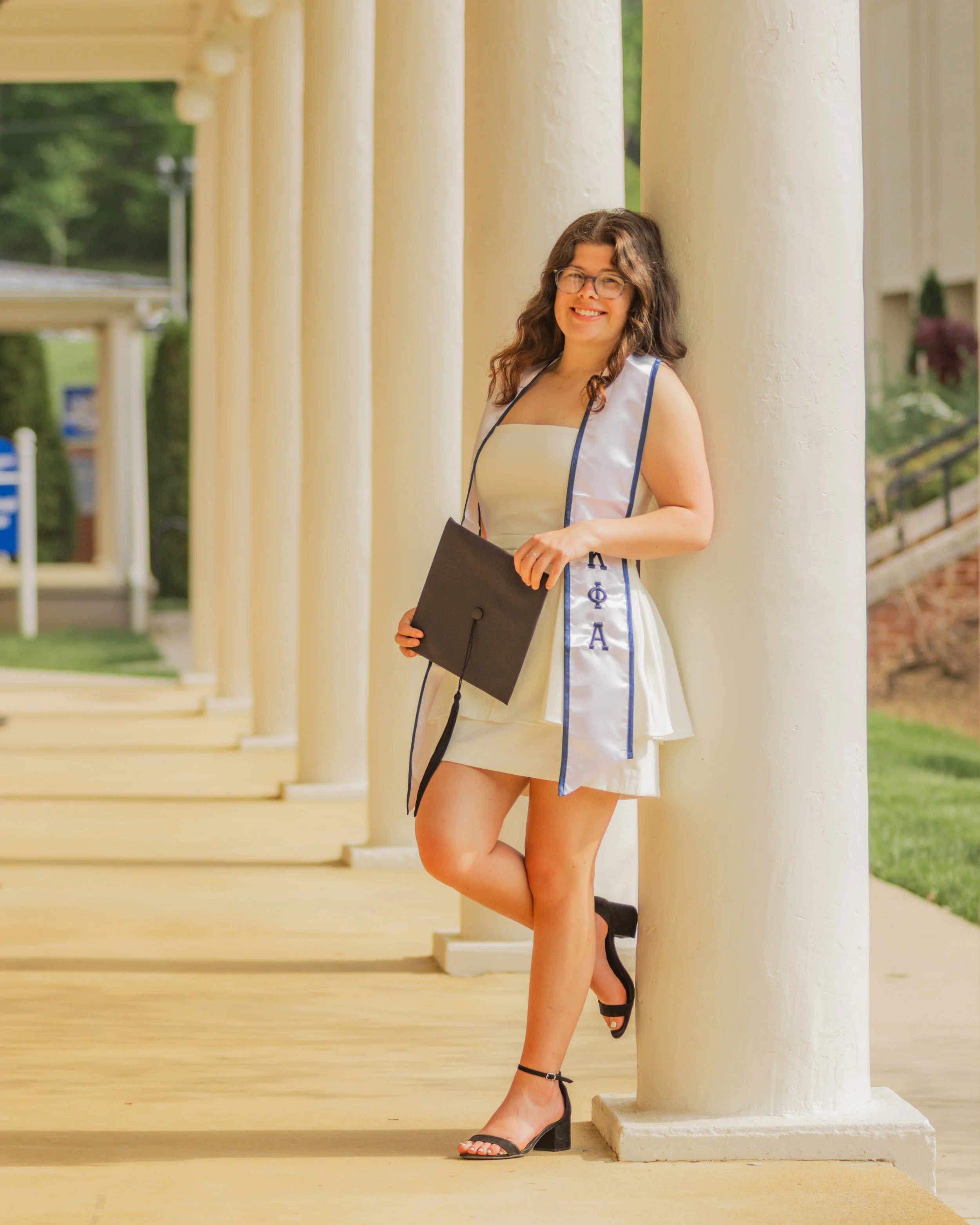 A woman in a white dress with a graduation stole, holding a mortarboard, standing outside near white columns, smiling.